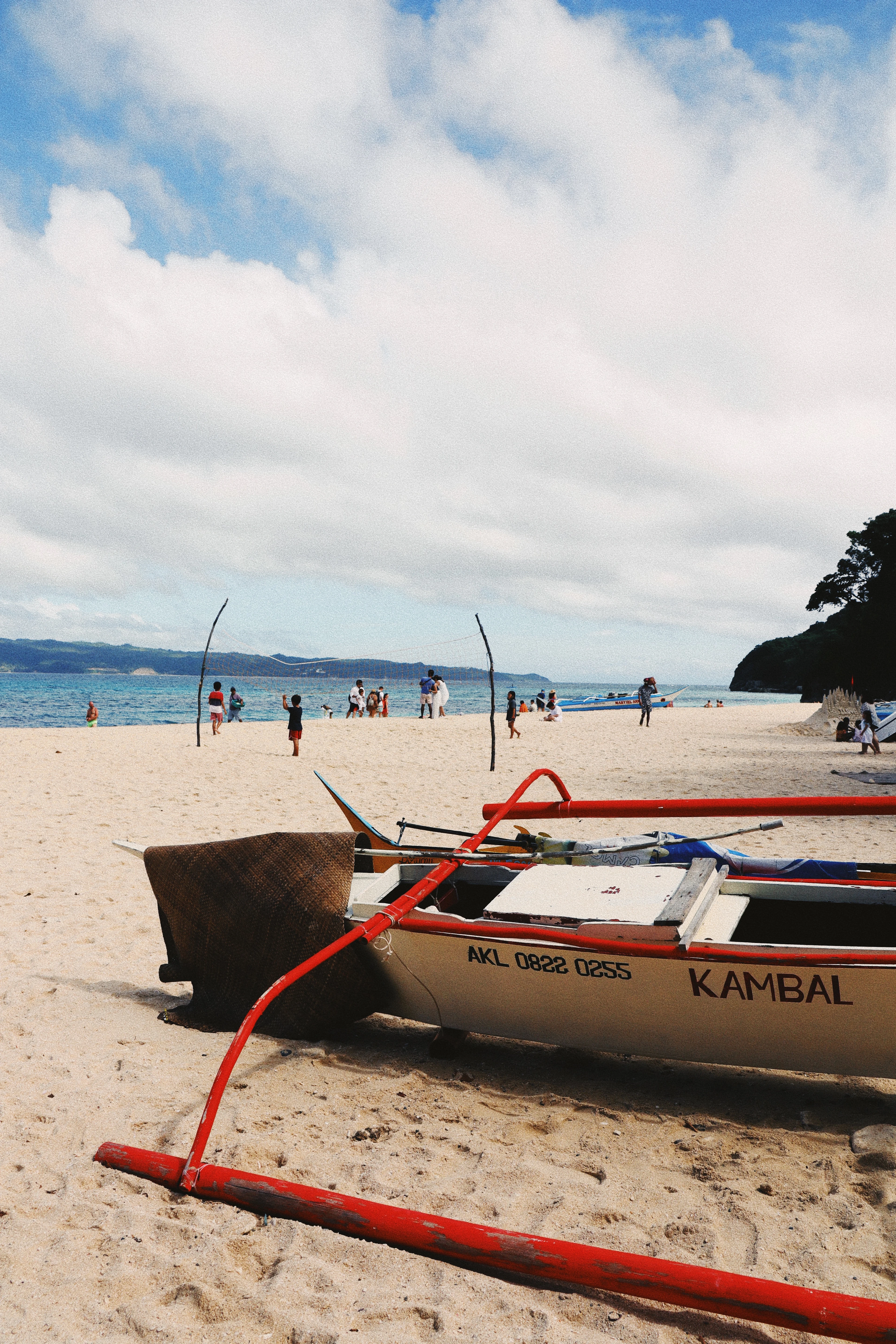 A red-trimmed outrigger boat rests on white sand as beachgoers wade in the turquoise shallows of Boracay.