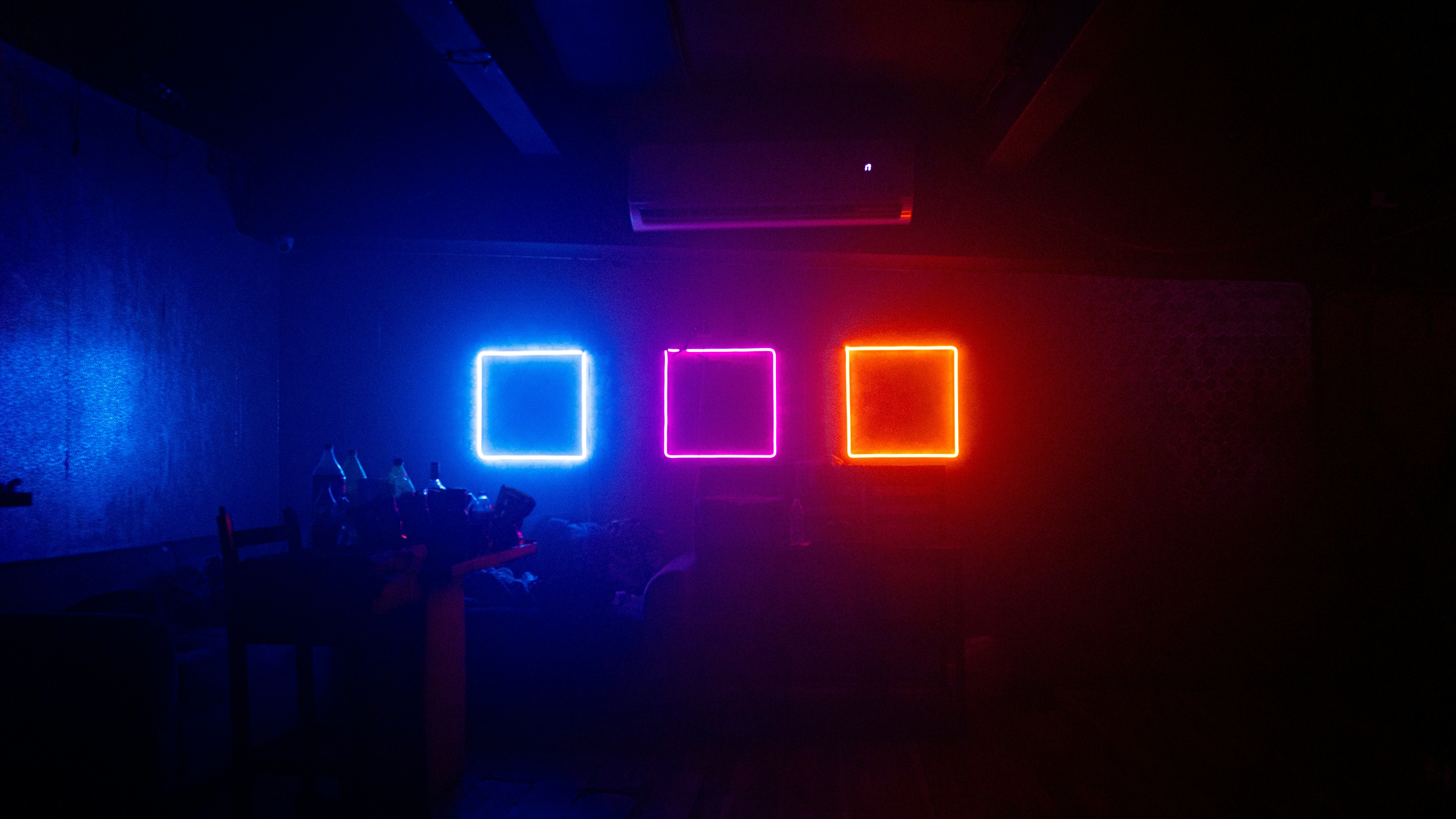 Tres cuadrados de neón en azul, magenta y naranja sobre una pared oscura.