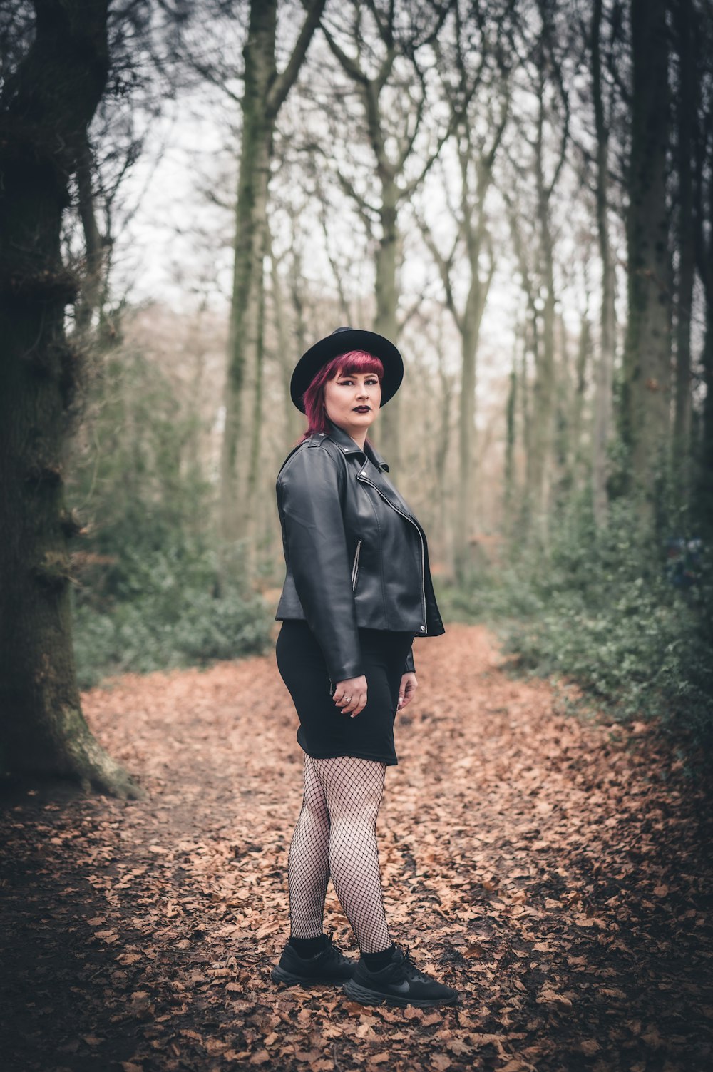 a woman in a black dress and hat standing in a forest