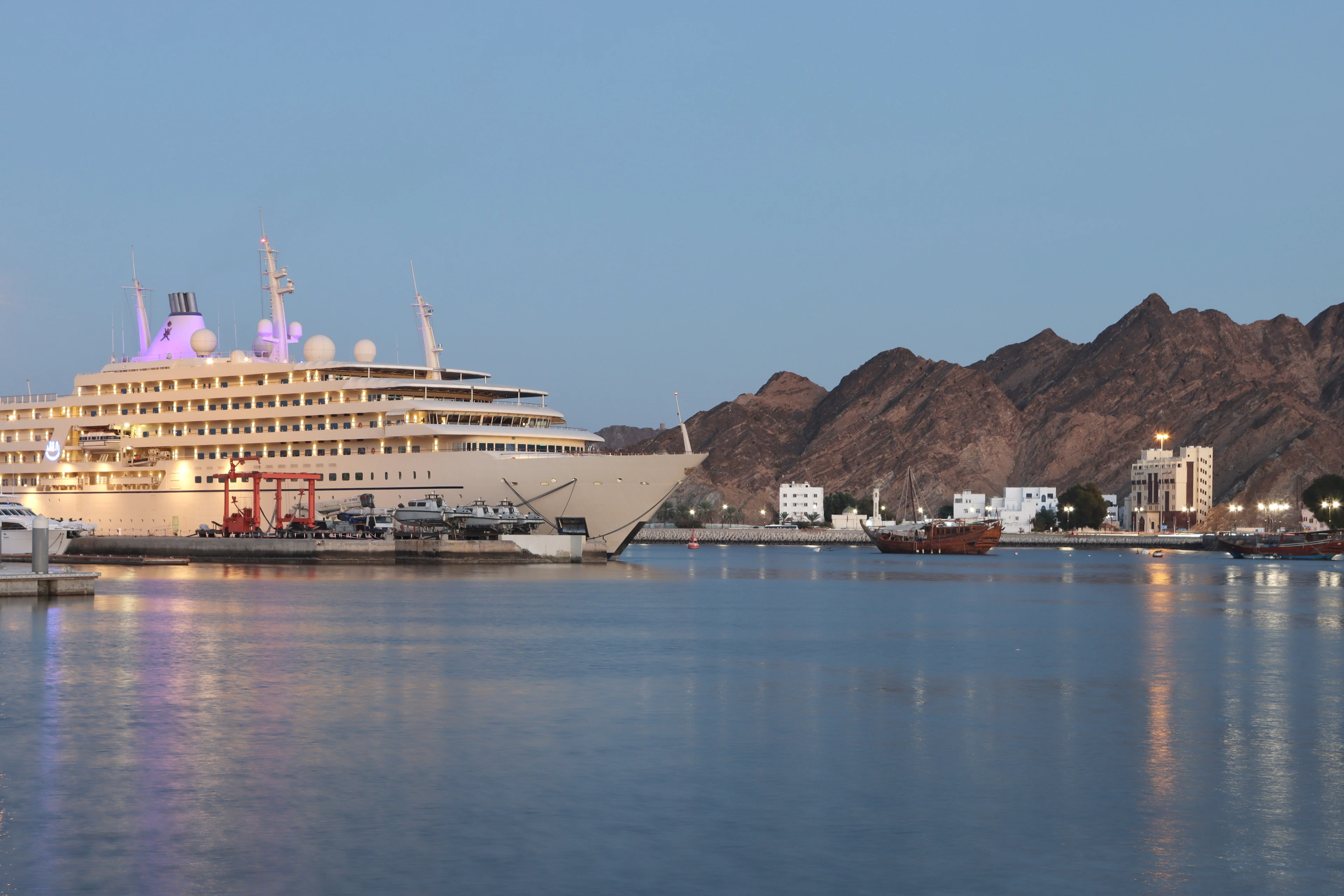 A large white cruise ship docked in Muscat's harbor at dusk, backed by dark mountains and calm water.