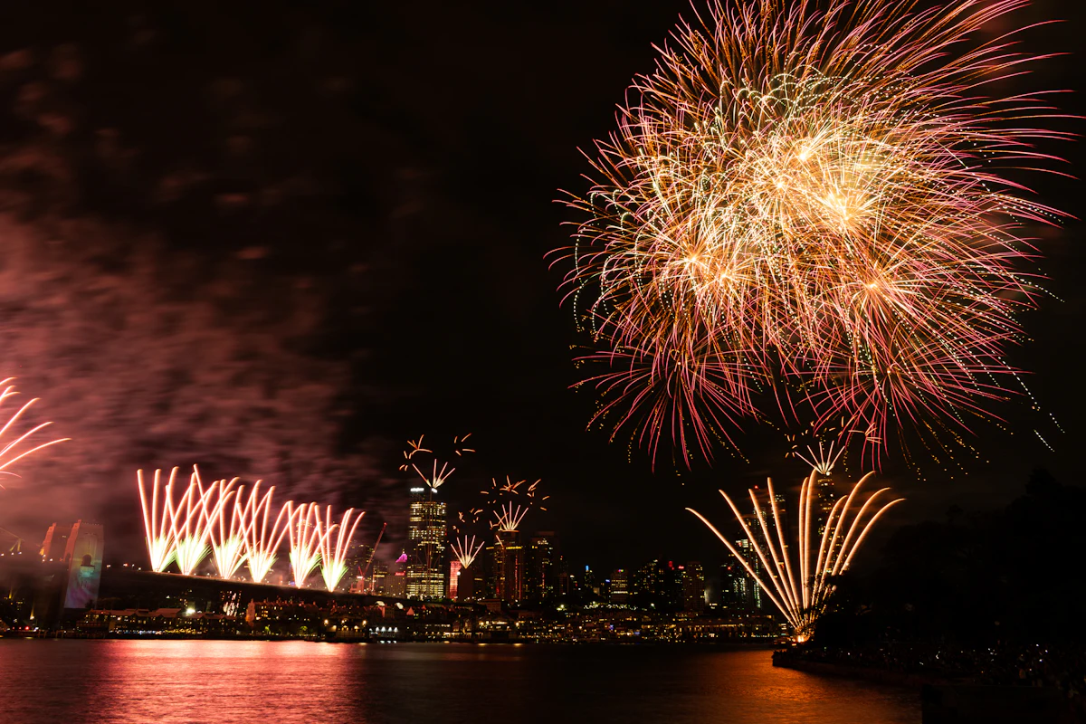 Sydney Harbour Bridge launching fireworks over the harbour with the city skyline and a shoreline crowd