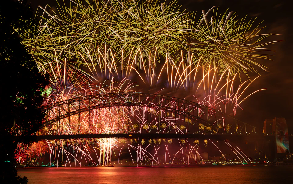A spectacular fireworks display over Sydney Harbour Bridge at night, with the city skyline illuminated and reflections in the harbour water