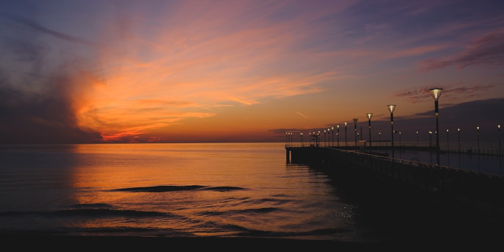 the sun is setting over the ocean with a pier in the foreground