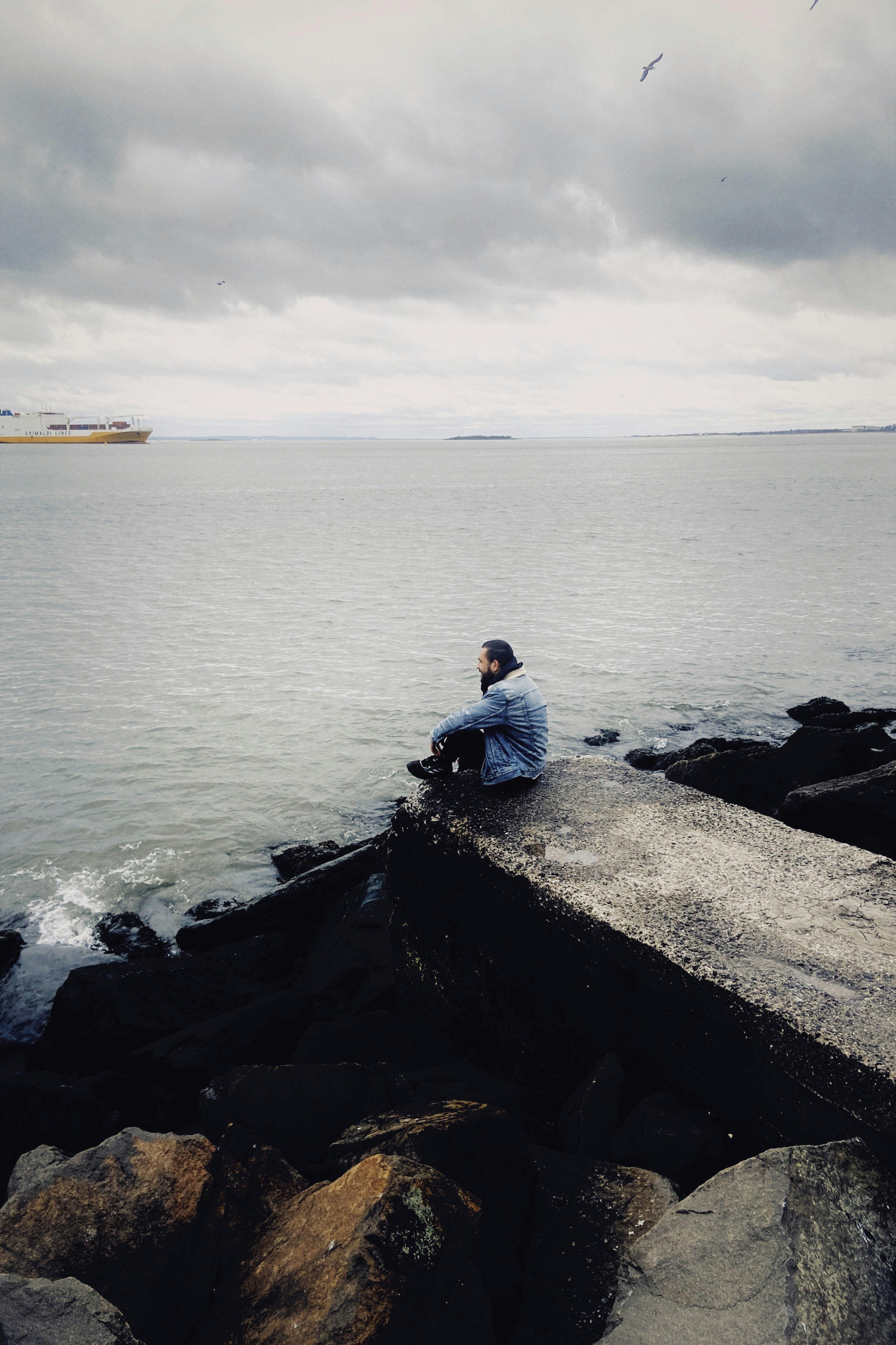 A man sitting on a rock next to the ocean photo – Free Bay ridge Image ...