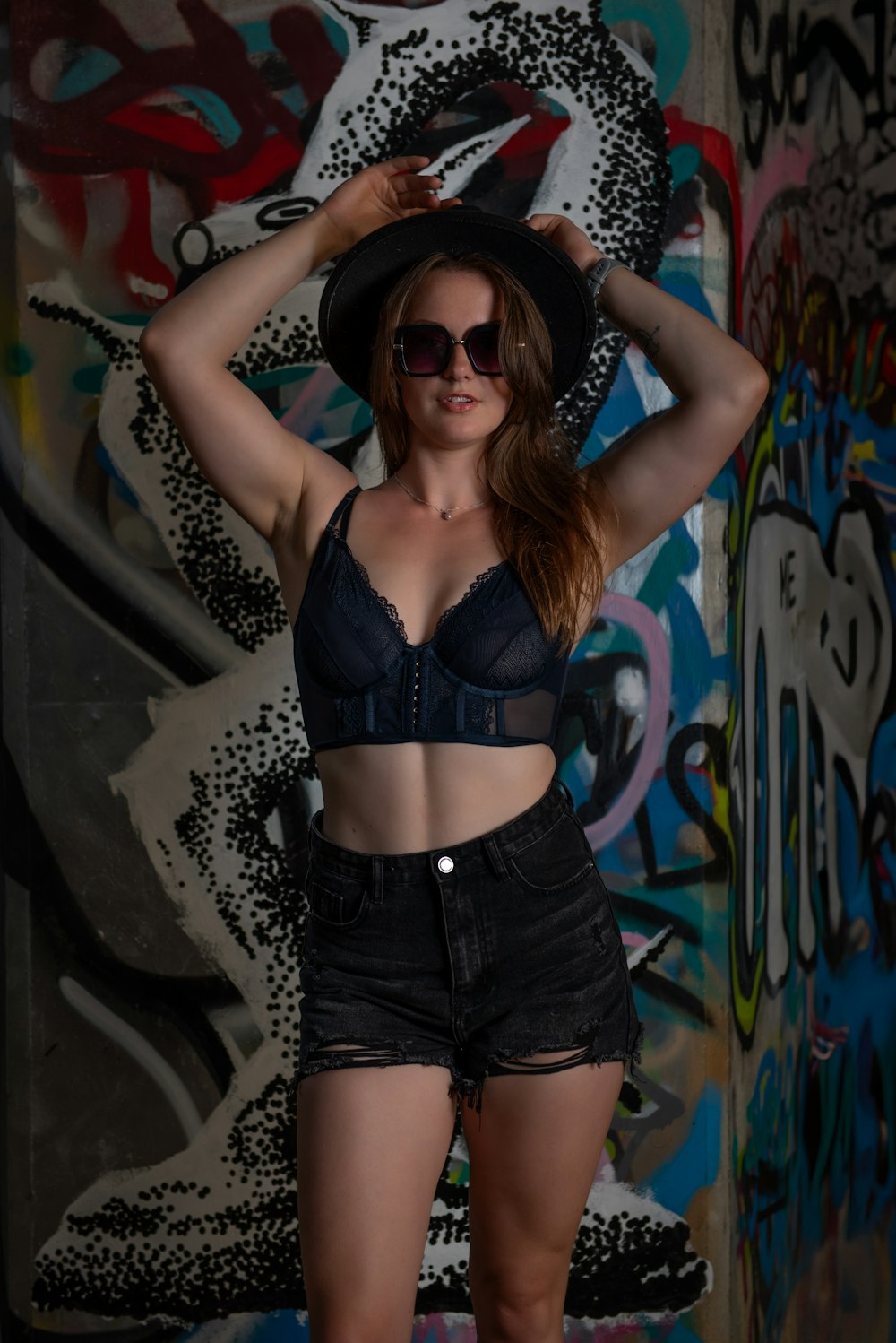 a woman wearing a hat and sunglasses posing in front of a wall with graffiti