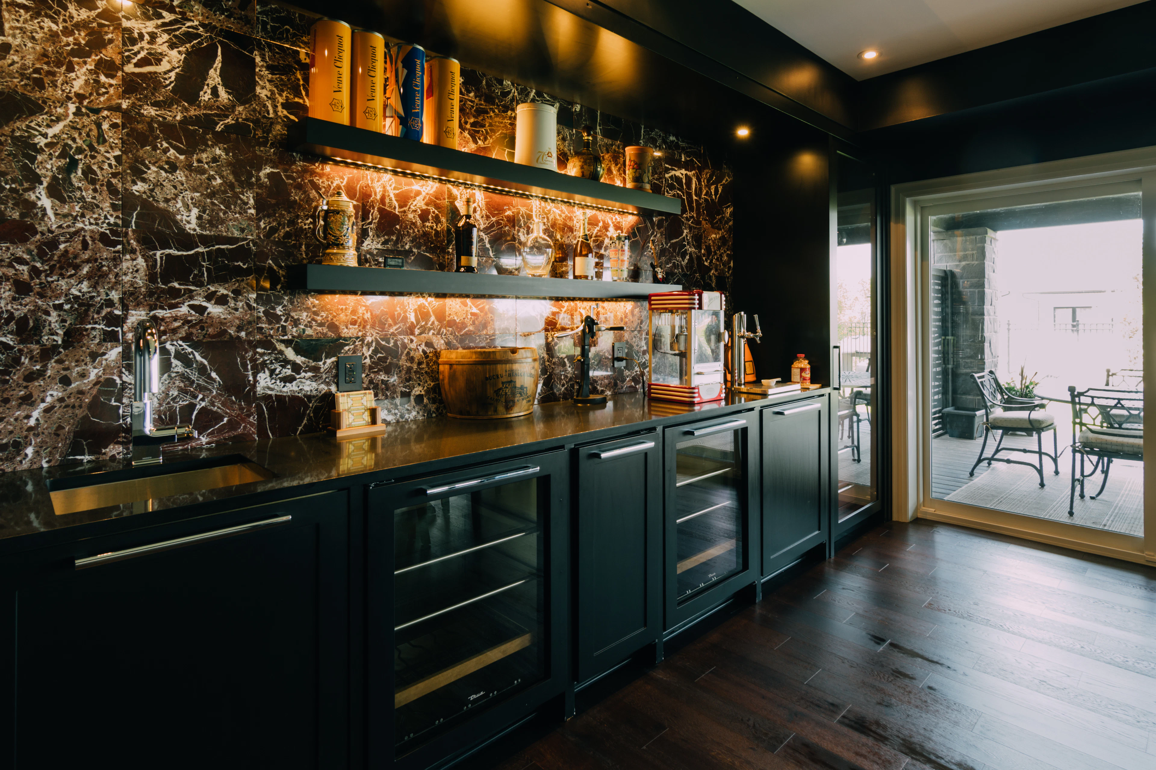 Dark green home bar with a stone backsplash and lit shelving