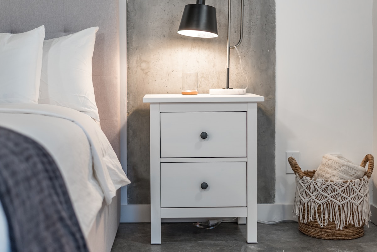 White nightstand with drawer and wicker basket beside apartment bed
