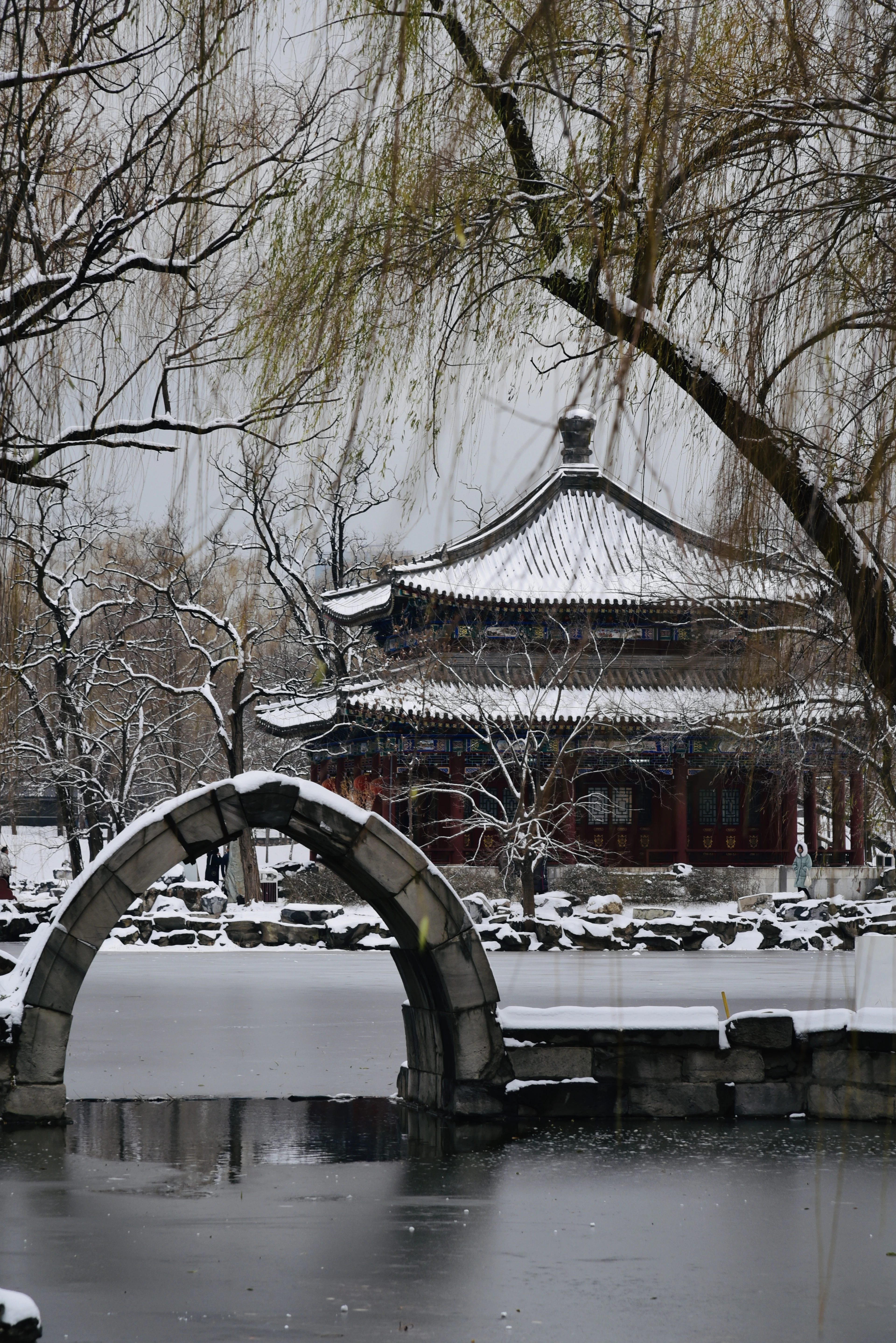 A snow-covered Chinese pavilion behind a stone moon-arch bridge and a half-frozen pond in a Beijing park, bare willow branches hanging down across the frame.