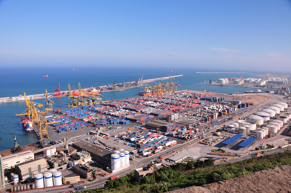 The image presents an expansive view of a bustling port city, teeming with activity and filled with various types of ships and cranes. The port is situated on a large body of water, which serves as a natural barrier for the city's development. The city itself is densely populated with buildings, including a large warehouse that appears to be a hub for maritime activities.

Numerous ships can be seen docked at the port, some larger than others and varying in color from red to blue. These ships are likely involved in different types of cargo transportation or maintenance tasks. The cranes present at the port indicate its capacity to handle large volumes of goods and materials.

The city itself is a mix of residential and commercial areas, with buildings housing various businesses and residences. The sky above the city is clear blue, suggesting good weather conditions for maritime activities.