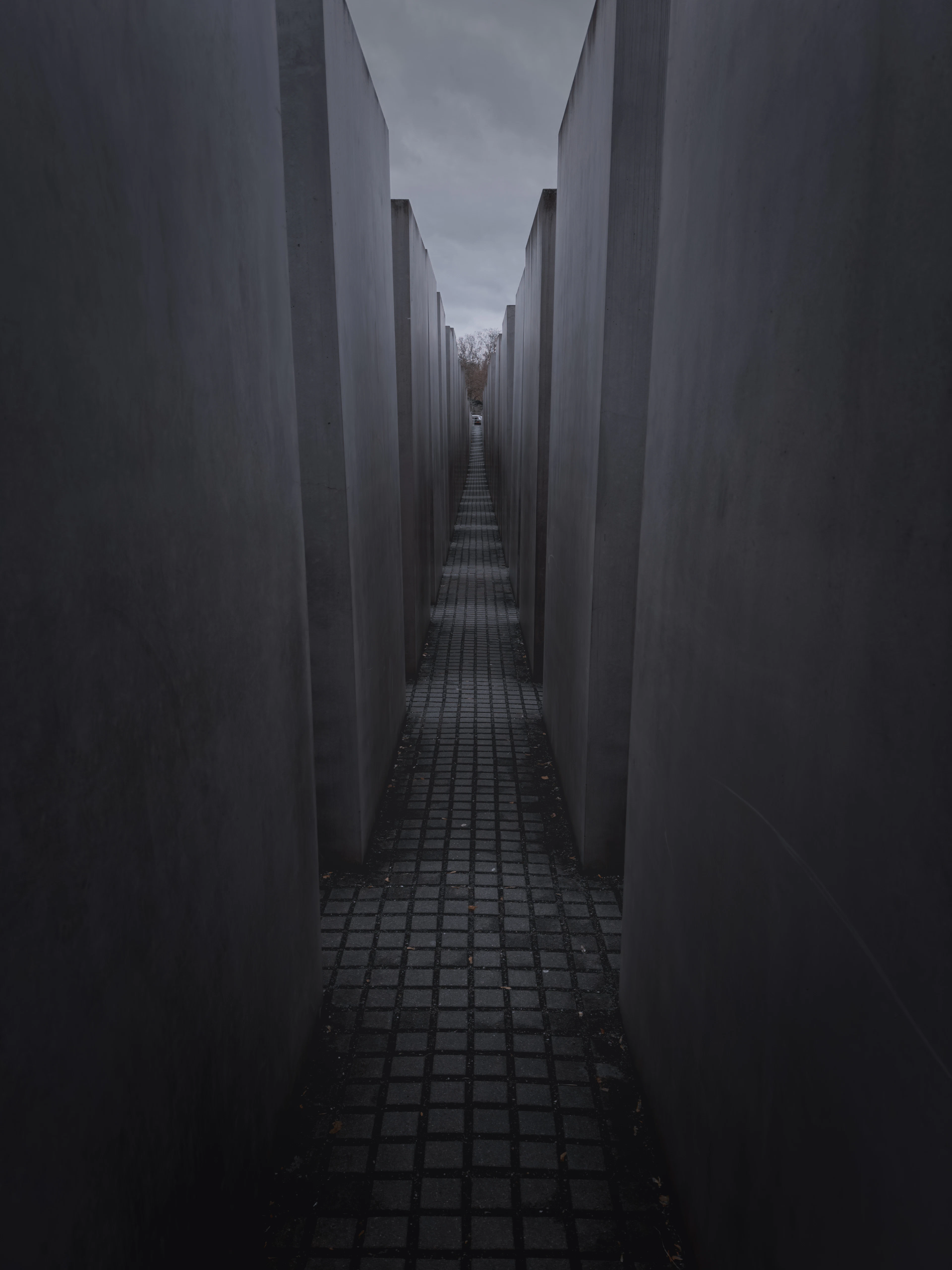 A narrow cobbled corridor running between the towering grey concrete stelae of Berlin's Memorial to the Murdered Jews of Europe, the ground dipping away under a pale overcast sky.