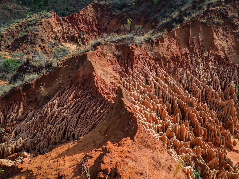The image depicts a rugged, red rock formation with numerous cracks and crevices that have been carved out over time by natural forces such as wind and water erosion. The formation is situated on a hillside, surrounded by vegetation including bushes and trees. The colors in the image are predominantly red from the rock formations and green from the vegetation, creating a striking contrast against the brown hues of the dirt ground.

The perspective of the photo is taken from below, looking up at the towering height of the formation, emphasizing its grandeur and the scale of nature's power that has shaped it over time.