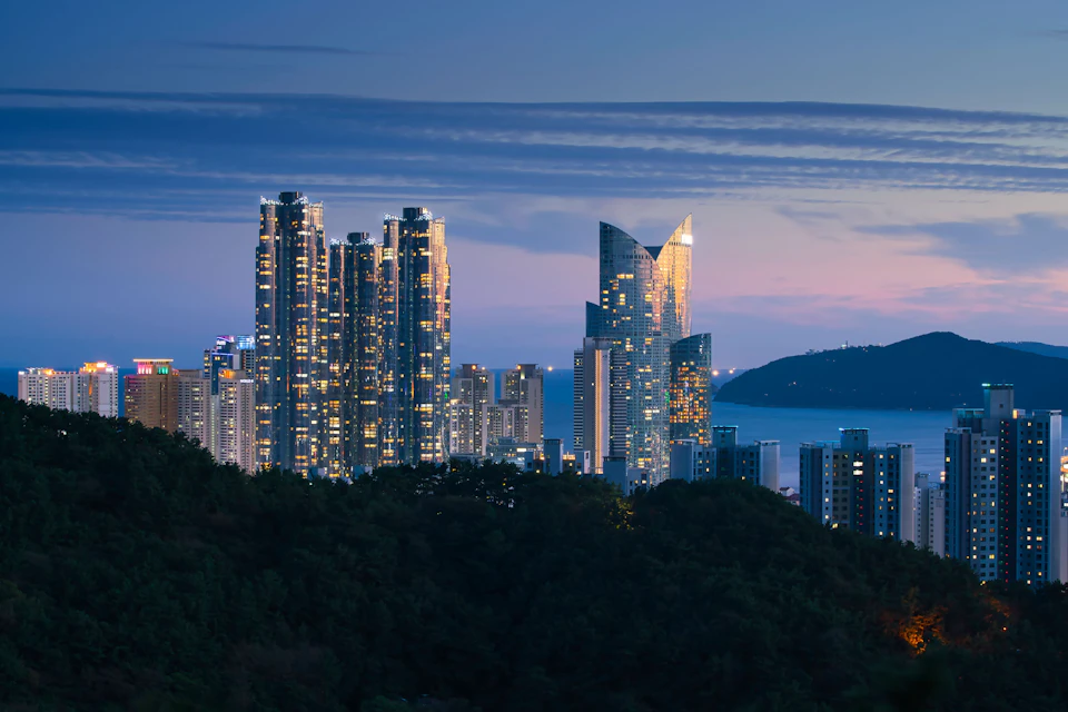 The image depicts a cityscape at dusk, with the sun setting over the horizon and casting a warm glow on the buildings below. The sky is painted in shades of blue, dotted with wispy clouds that add depth to the scene. The city skyline features tall buildings of varying heights, some of which are illuminated by artificial lights, creating a vibrant contrast against the darkening sky. In the distance, a body of water can be seen, adding a serene element to the urban landscape.