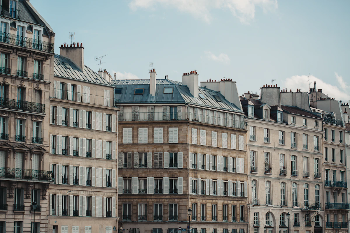 Une rangée d'immeubles anciens aux façades claires, avec des volets, des balcons en fer forgé et des toits en zinc, sous un ciel légèrement nuageux