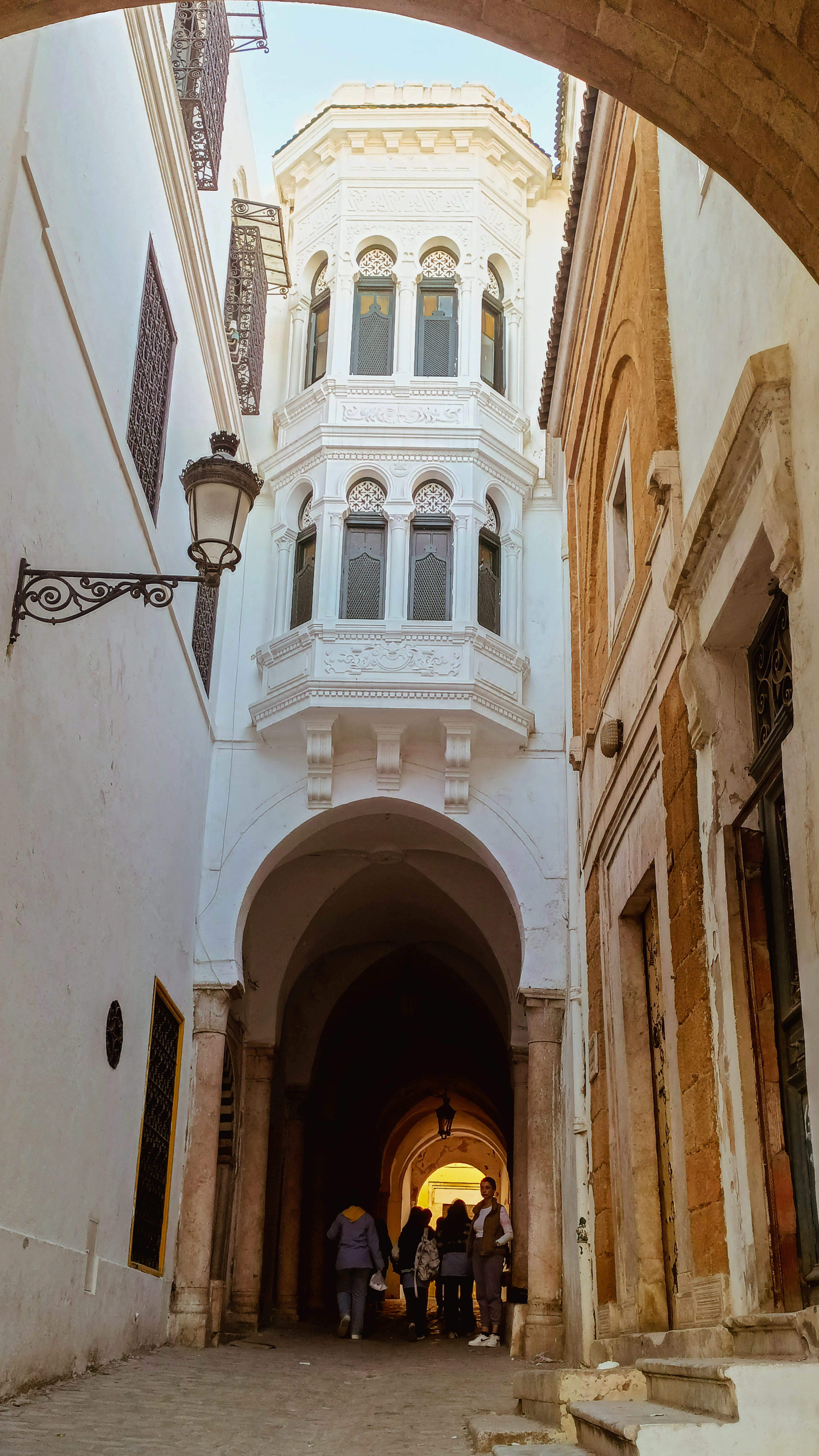 A white carved oriel window overhanging a vaulted stone passage in the Tunis medina, a handful of people walking through toward the sunlit lane at the far end.