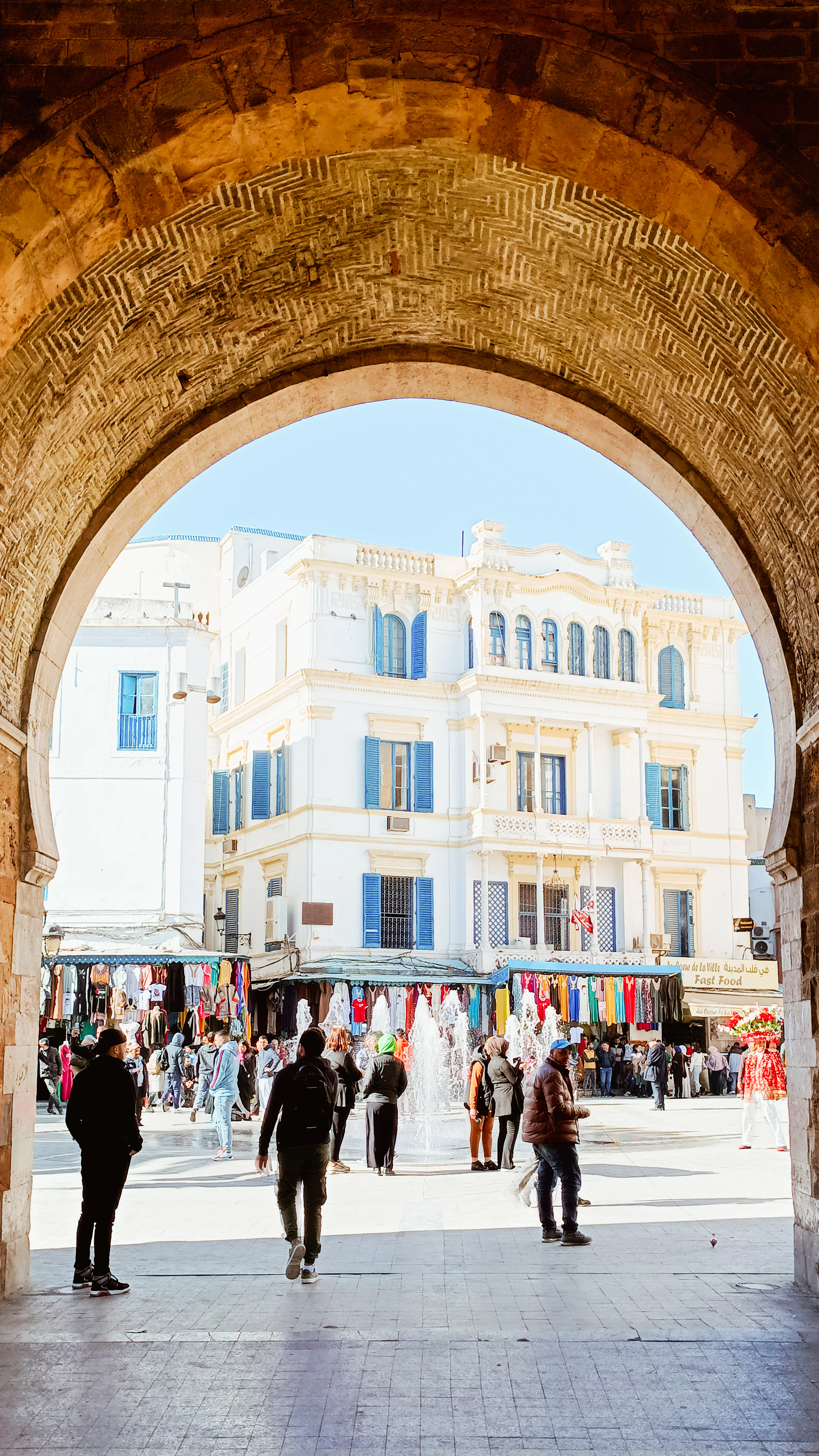Looking out through the great stone arch of Bab El Bhar in Tunis to the white colonial facades, blue shutters, clothing stalls and fountain of Place de la Victoire beyond.