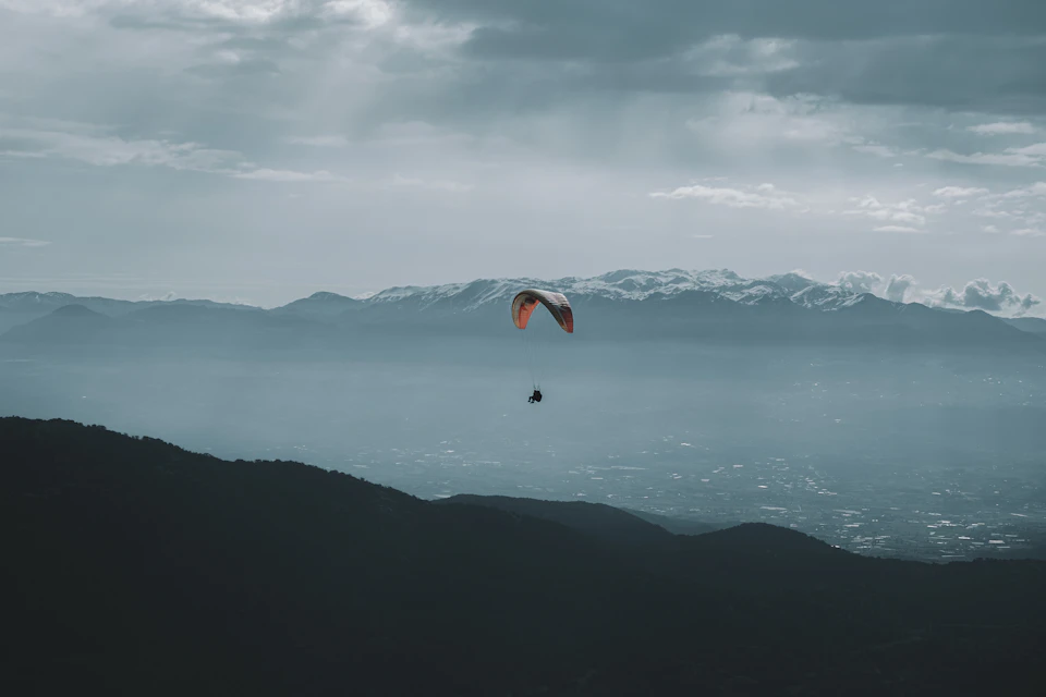 Person paraglides over snowy mountain range with open parachute and soaring through cloudy sky