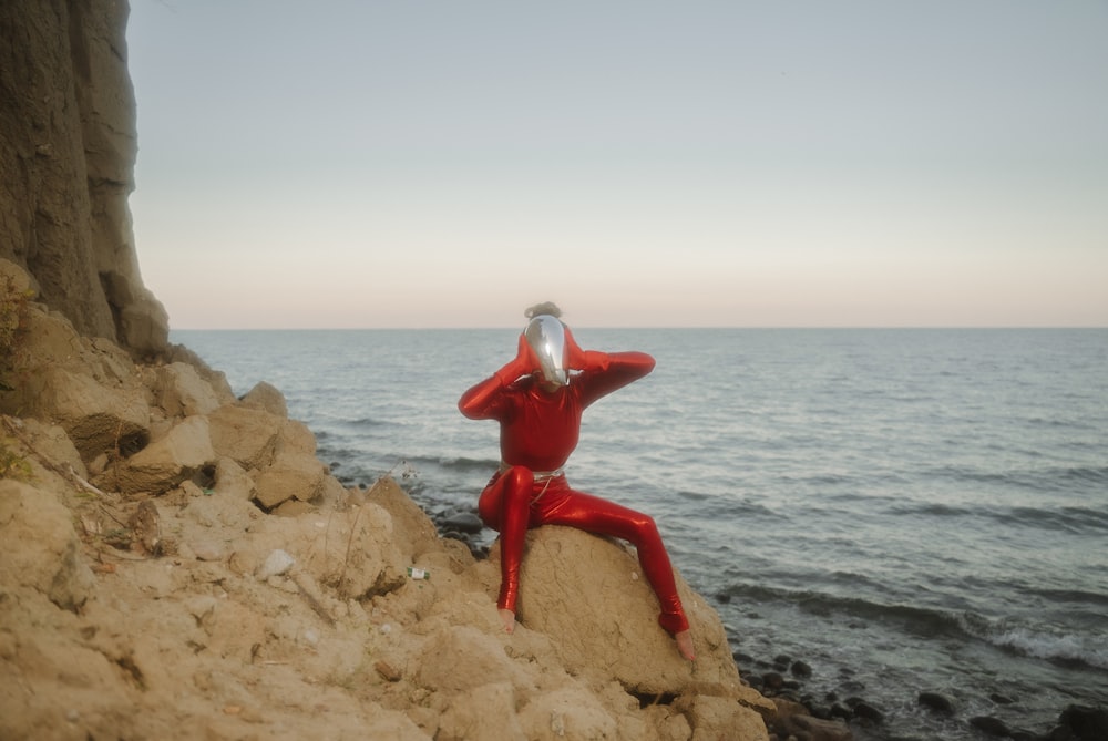 a person in a red suit sitting on a rock near the ocean