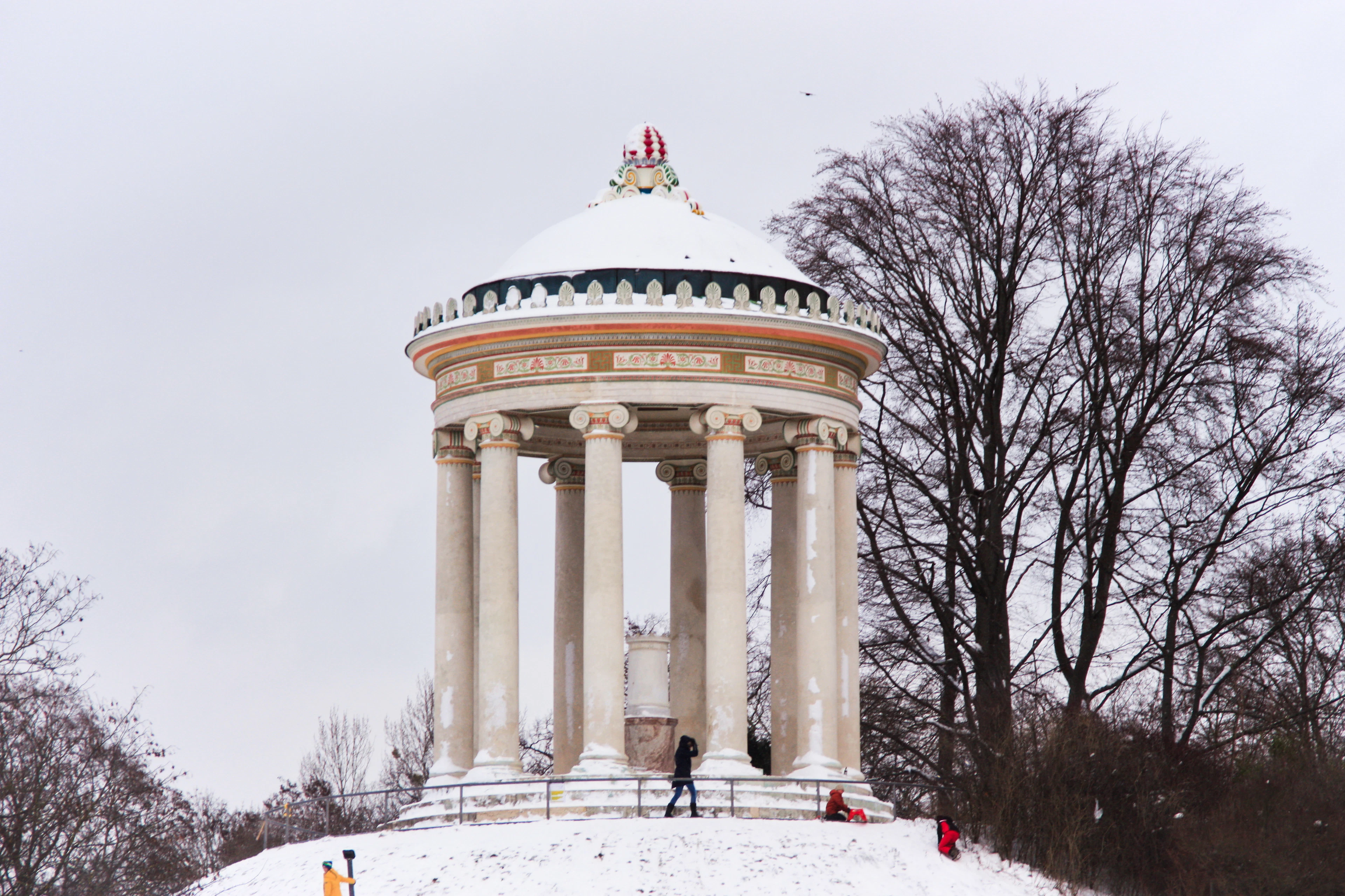 The Monopteros temple on its snow-covered mound in Munich's English Garden in midwinter, bare beeches behind it and a handful of walkers climbing the white slope