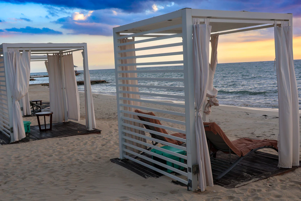 The image depicts a serene beach scene with two white gazebos, each featuring a white canopy and a green roof, situated on the sandy shoreline. The gazebos are positioned side by side, creating an inviting space for relaxation. In front of one of the gazebos, there is a table accompanied by two chairs, suggesting that this area could be used for dining or enjoying the view.

The beach itself is expansive and stretches out to meet the ocean at the horizon. The sky above is painted with hues of orange and pink, indicating that it's either sunset or sunrise. The water in the distance appears calm, adding to the tranquil atmosphere of the scene.