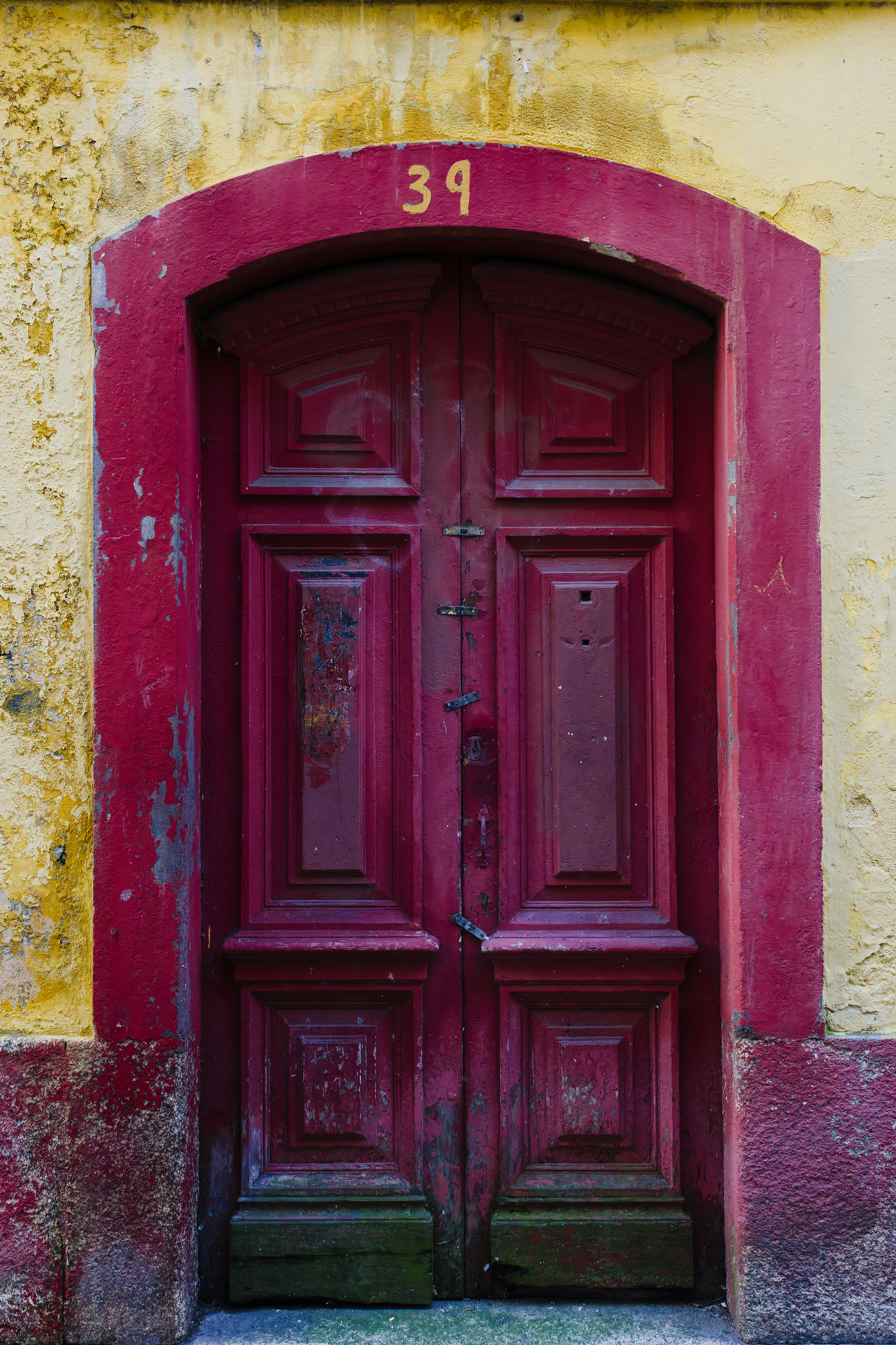 Red Wooden Door With Number Thirty Nine on a Yellow Wall in a European City - A red wooden door with ornate carvings and a number thirty nine painted in yellow above it, is set into a yellow wall with a faded red trim. The wall and trim show signs of age and weathering.