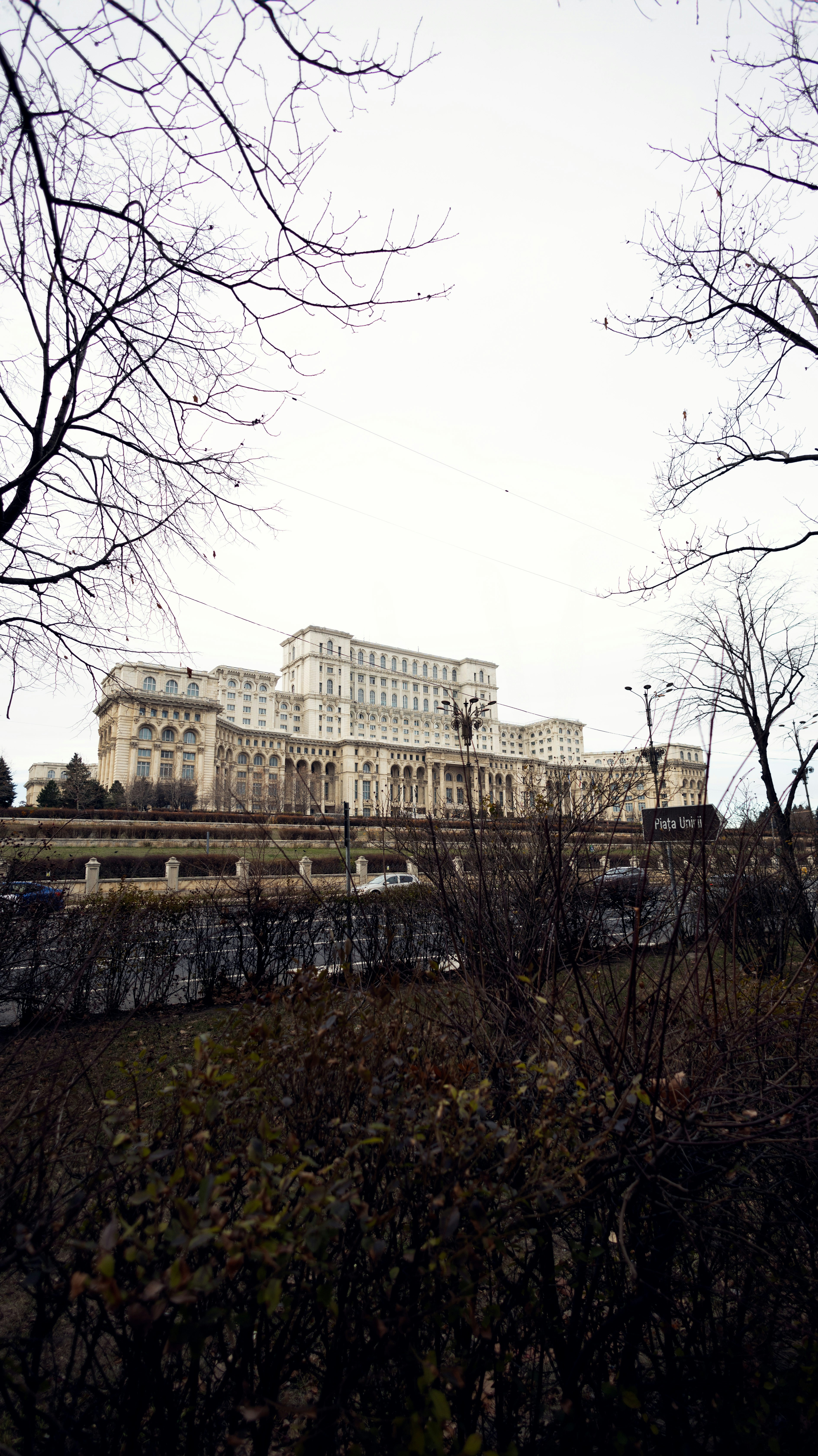 Cloudy day at the grand Palace of the Parliament in Bucharest, Romania, captured on January 8, 2024. Cars parked in front, bare trees frame the view.