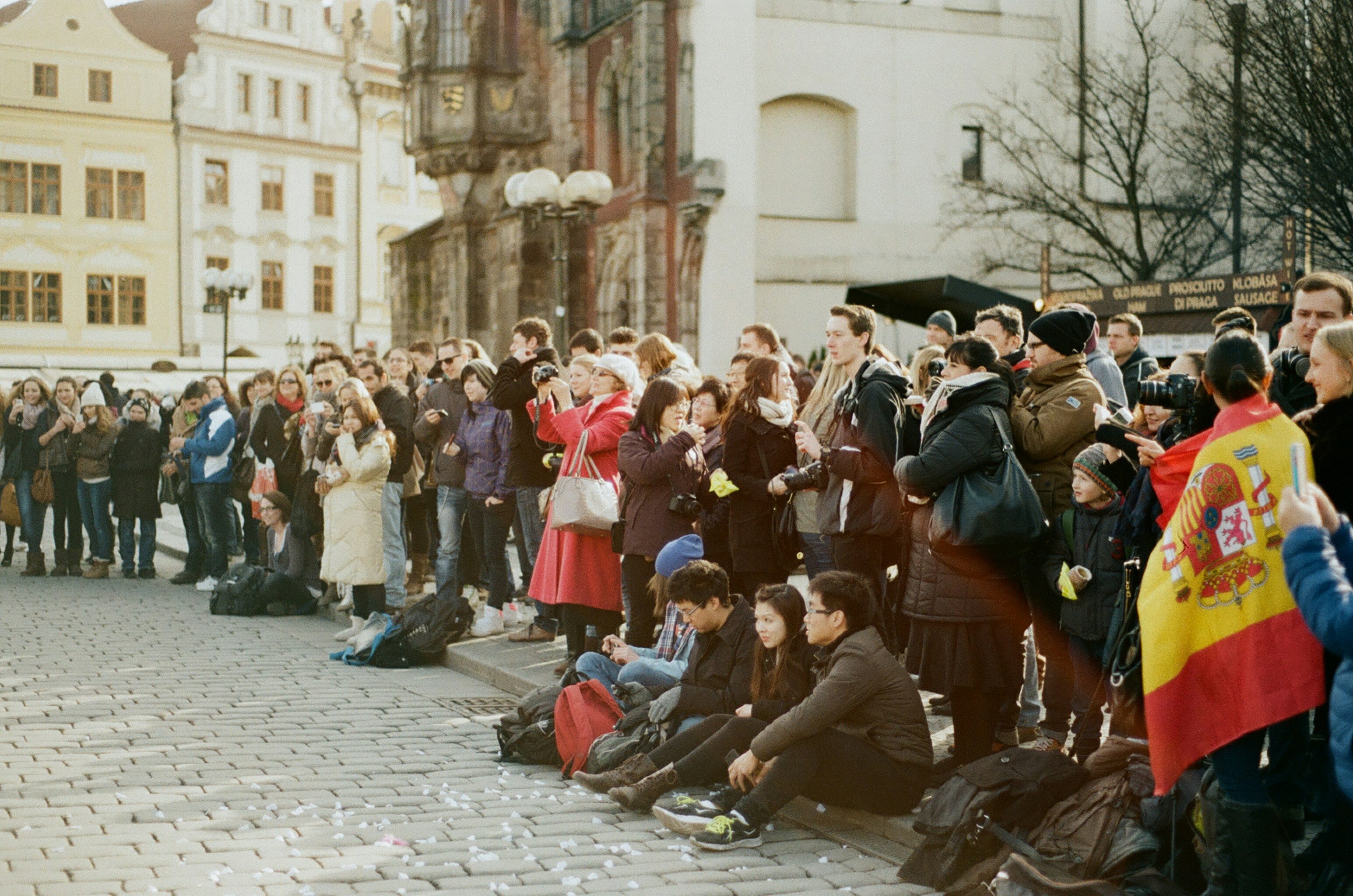 a crowd of people standing and sitting in front of a building, People sit and stand in Prague watching a flash mob.