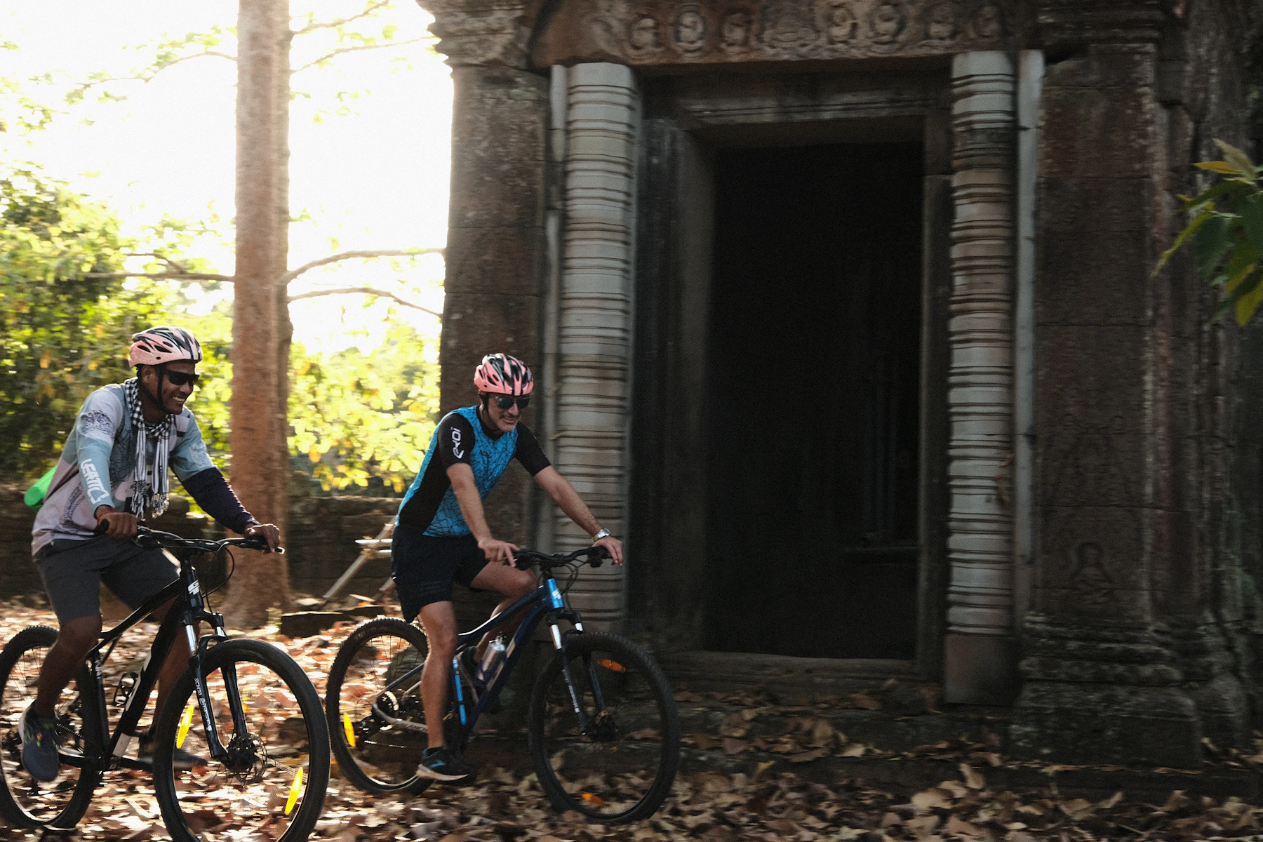 Two helmeted travelers cycling beside an ancient Khmer temple doorway in the Cambodian forest