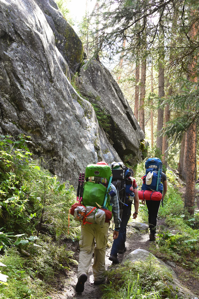 Hikers with backpacks walking in single file along a narrow trail beside a rock face