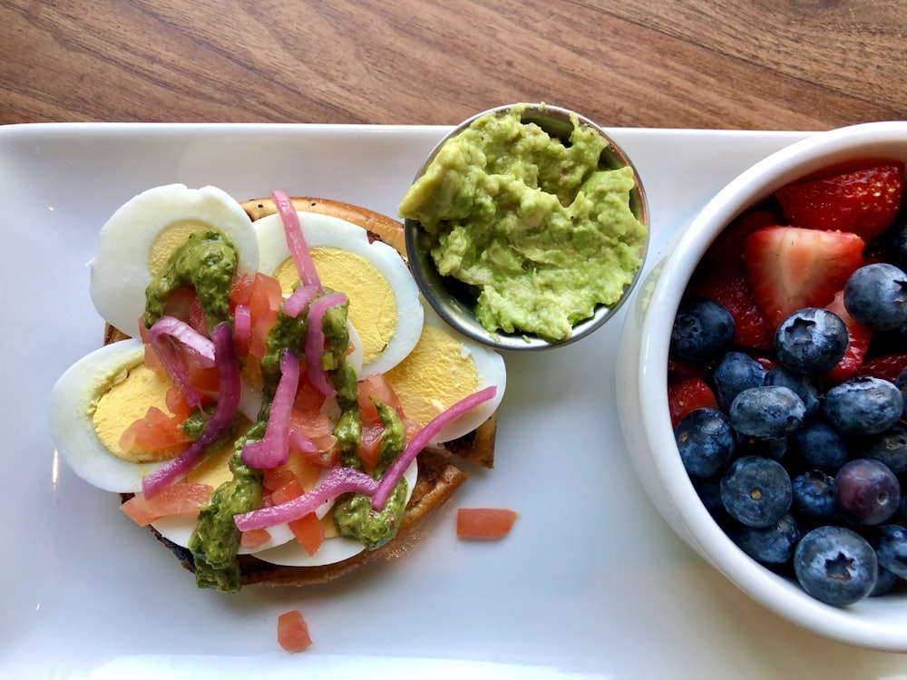 a white plate topped with a sandwich next to a bowl of fruit