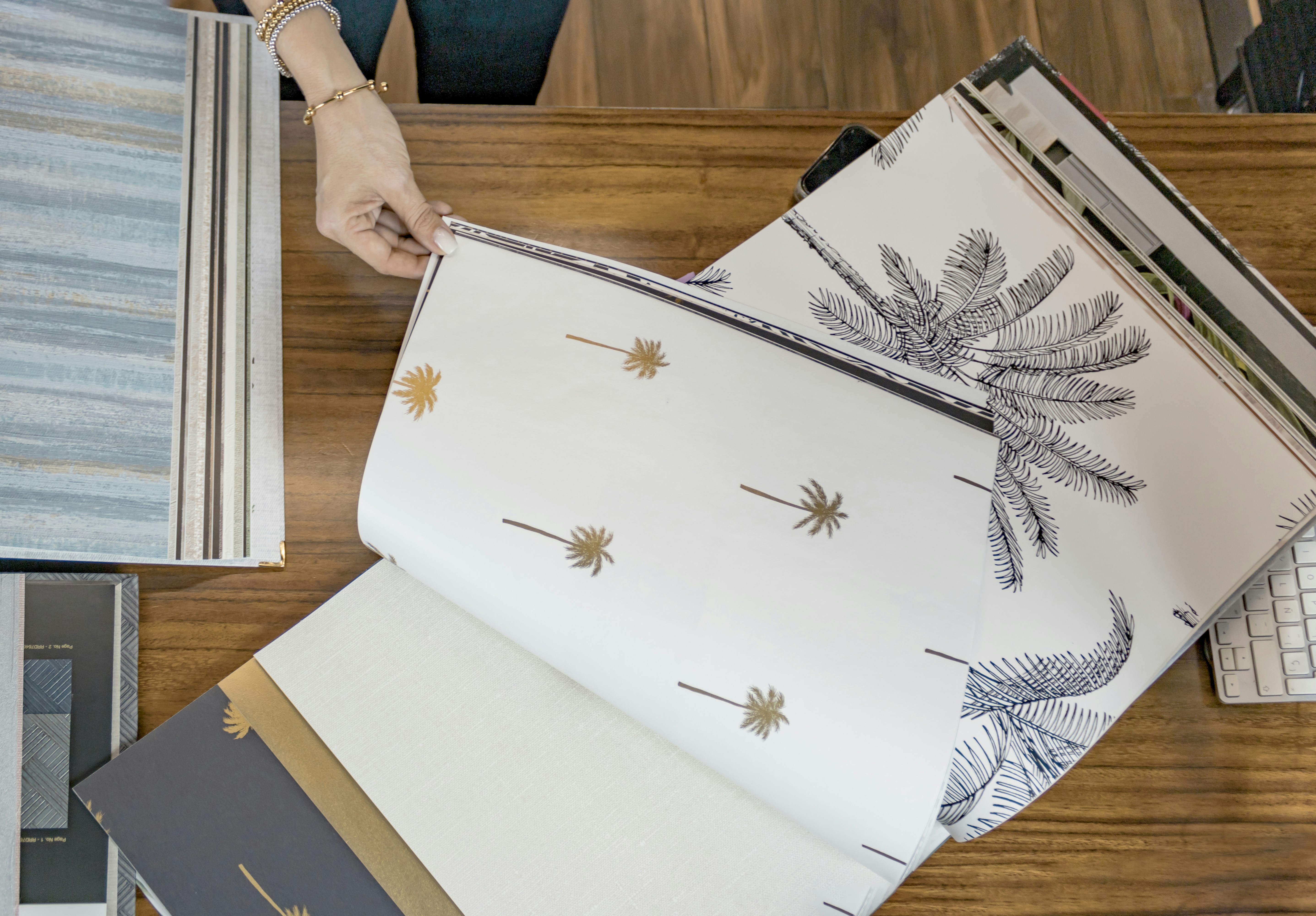 Interior designer flipping through wallpaper sample books with palm tree patterns on a wooden desk