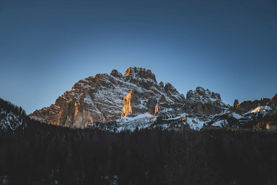 The image depicts a majestic mountain range with snow-capped peaks against a clear blue sky. The mountains, bathed in sunlight, stand tall and proud, their rocky surfaces gleaming under the warm rays. The perspective is from below, looking up at these towering natural wonders. In the foreground, there are trees that add depth to the scene, providing a sense of scale and highlighting the grandeur of the mountains.