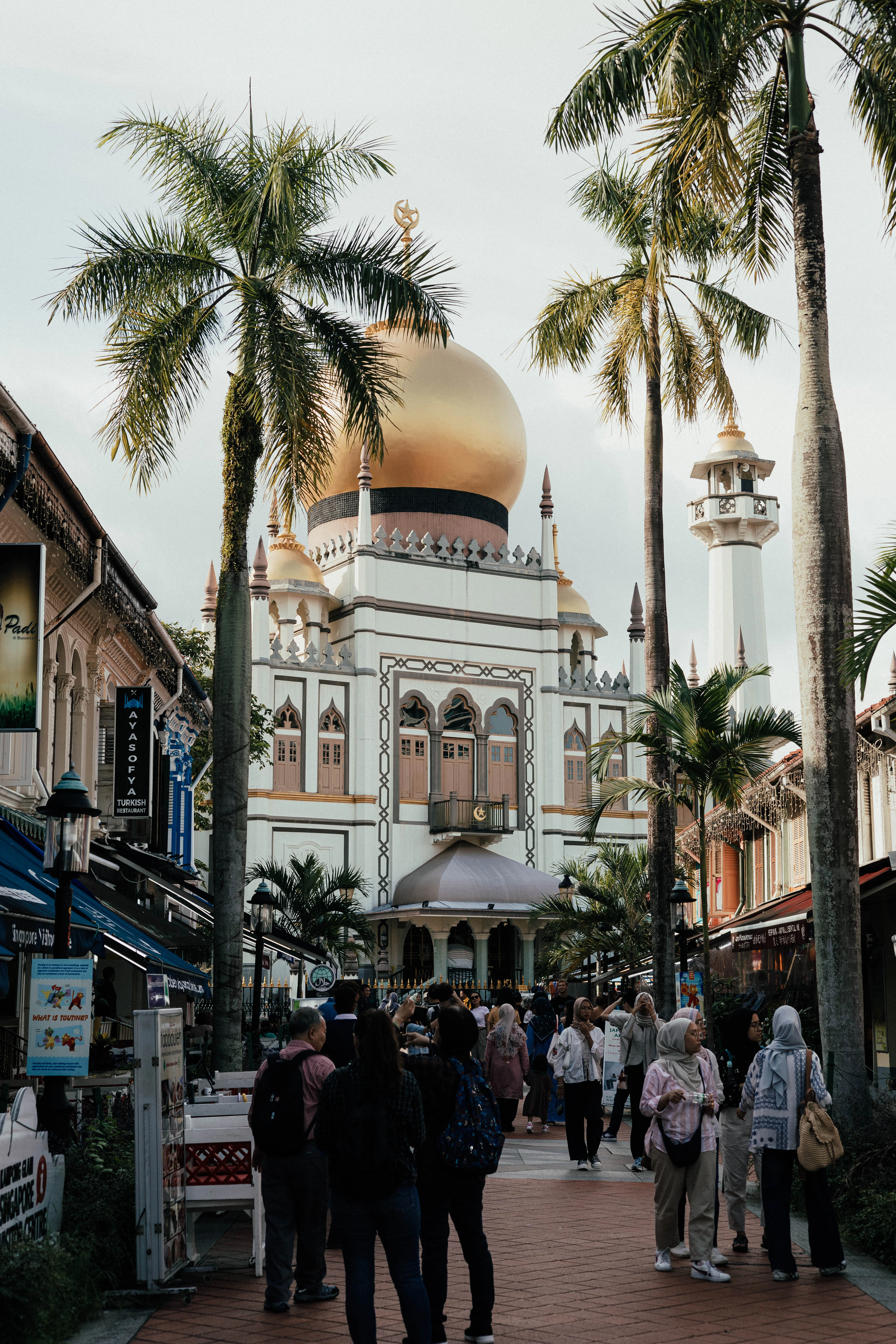 The golden dome and white minaret of Masjid Sultan rising above the palm-lined pedestrian stretch of Bussorah Street in Singapore's Kampong Glam, busy with visitors in the late afternoon.