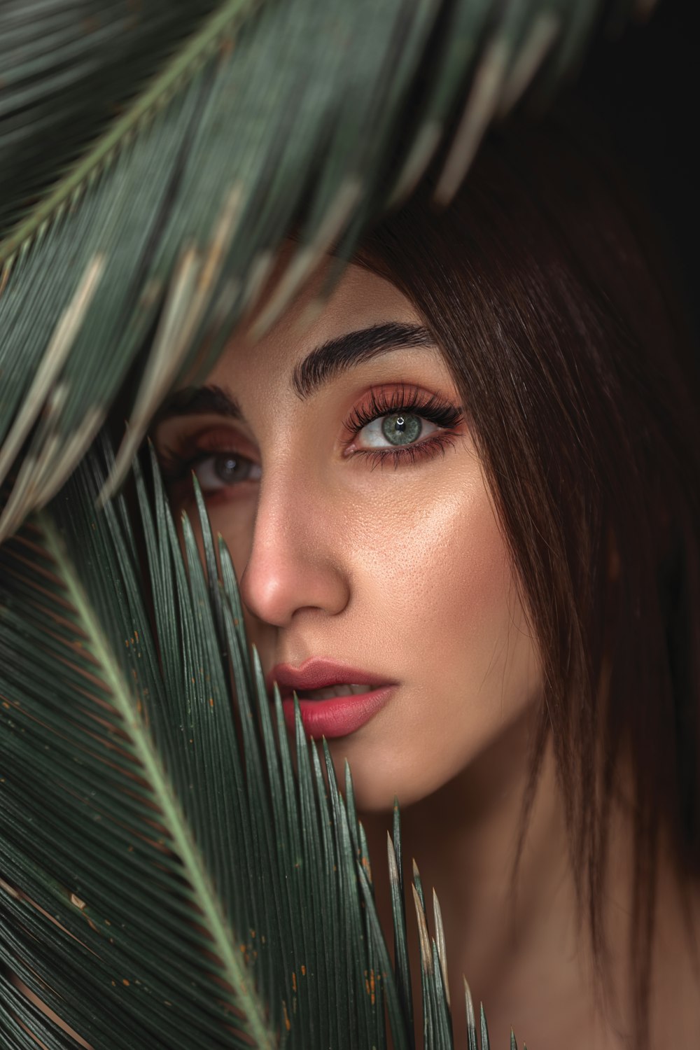 a woman with blue eyes hiding behind a palm leaf