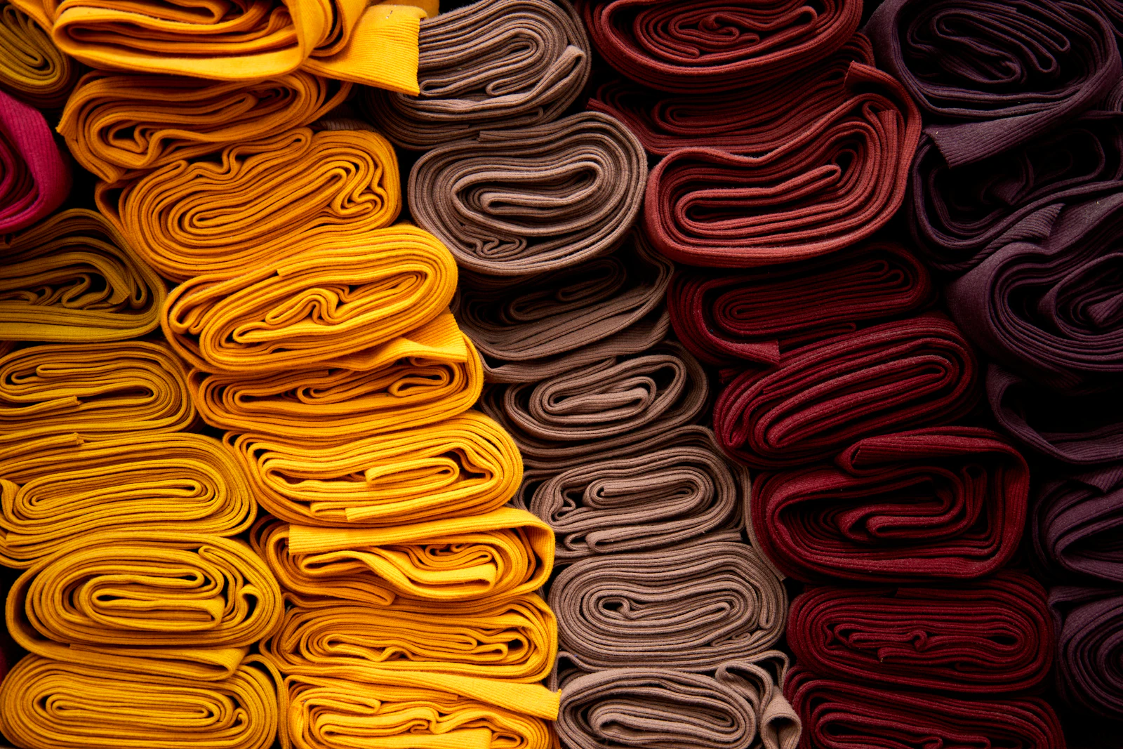 Neatly folded stacks of ribbed knit jersey fabric in yellow, tan, and deep red tones sorted by color
