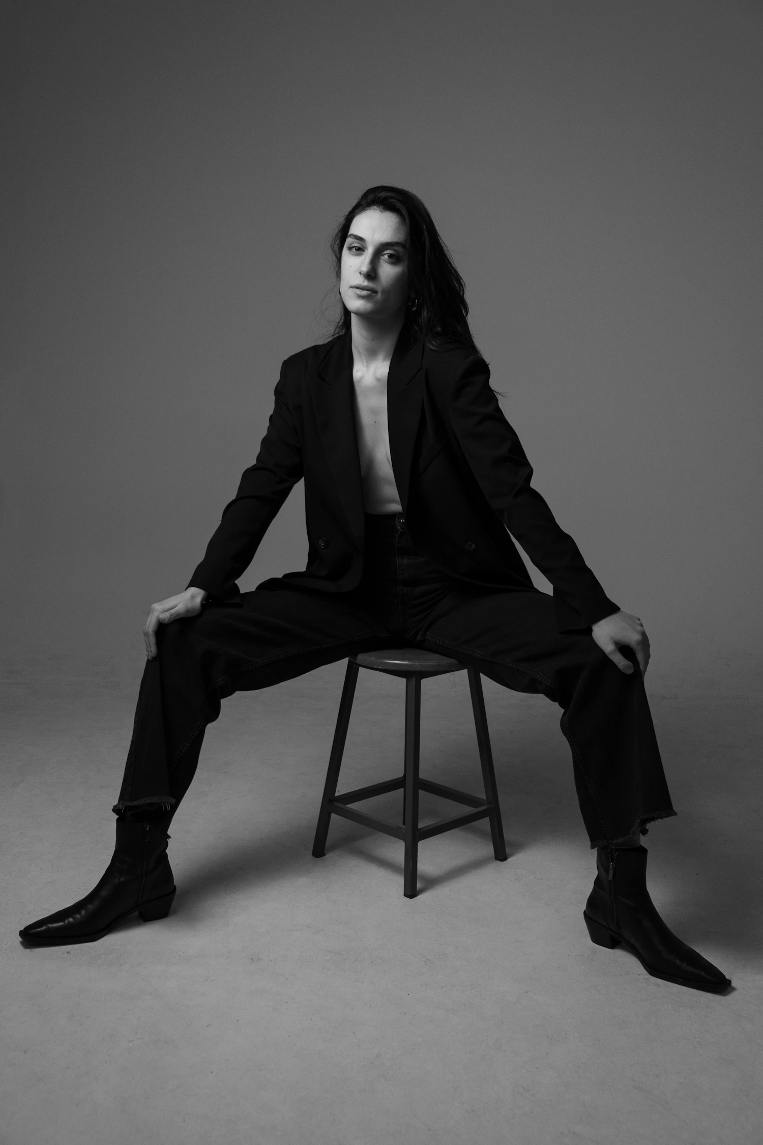 A black two-piece suit worn seated on a stool in a dark studio