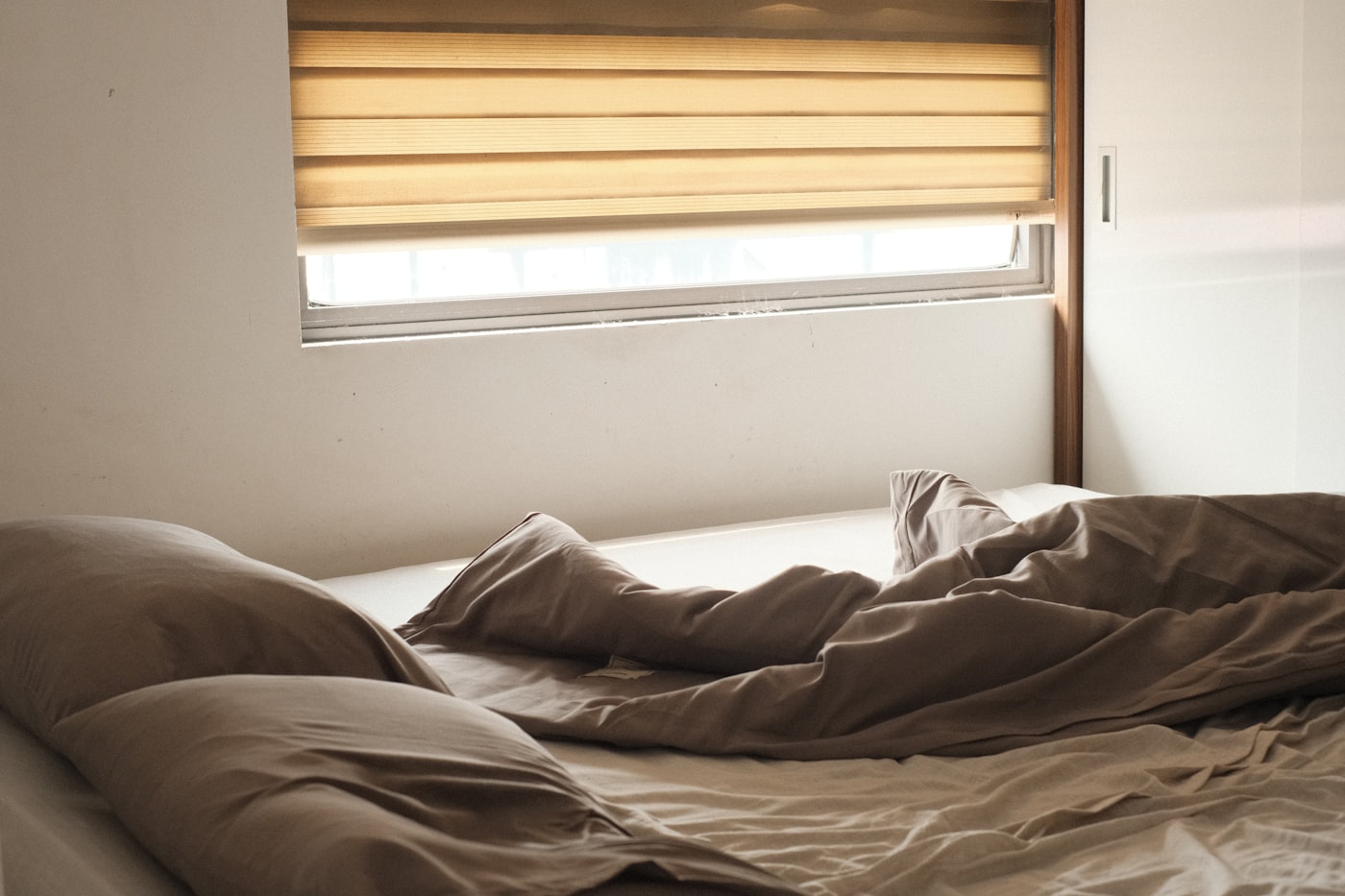 Minimalist apartment bedroom bathed in warm morning light with soft tan and warm brown linen bedding, amber cellular shade casting golden glow over a cozy earth tone palette