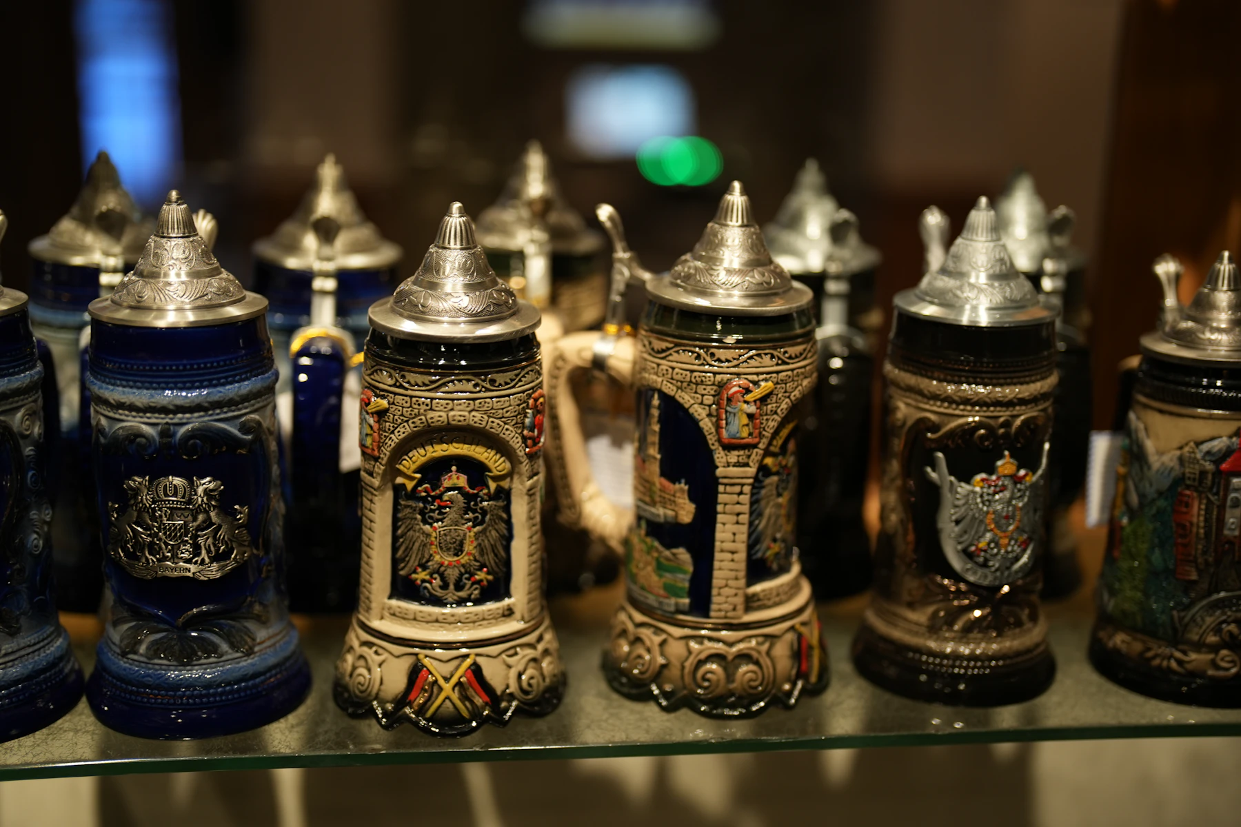 A row of relief-decorated beer steins with pointed pewter lids standing on a glass shelf