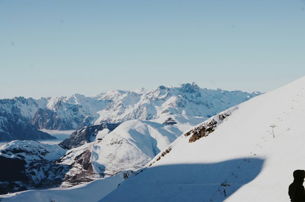A snowy ridge with a lift tower and ski tracks above Les Deux Alpes, looking across the Romanche valley to a wall of peaks