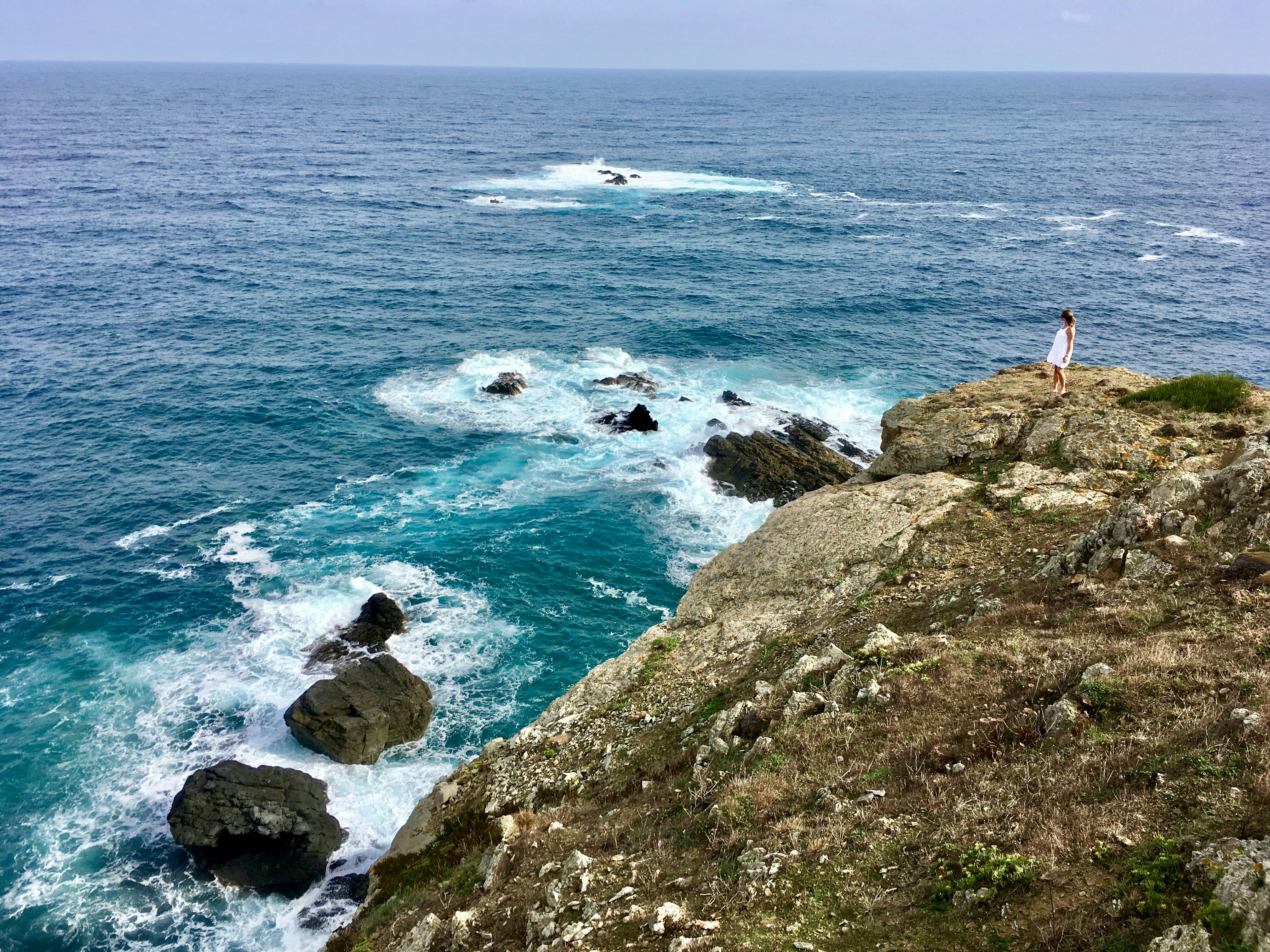 a person standing on top of a cliff next to the ocean, Woman in Menorca Island in White Dress. Shot with iPhone.