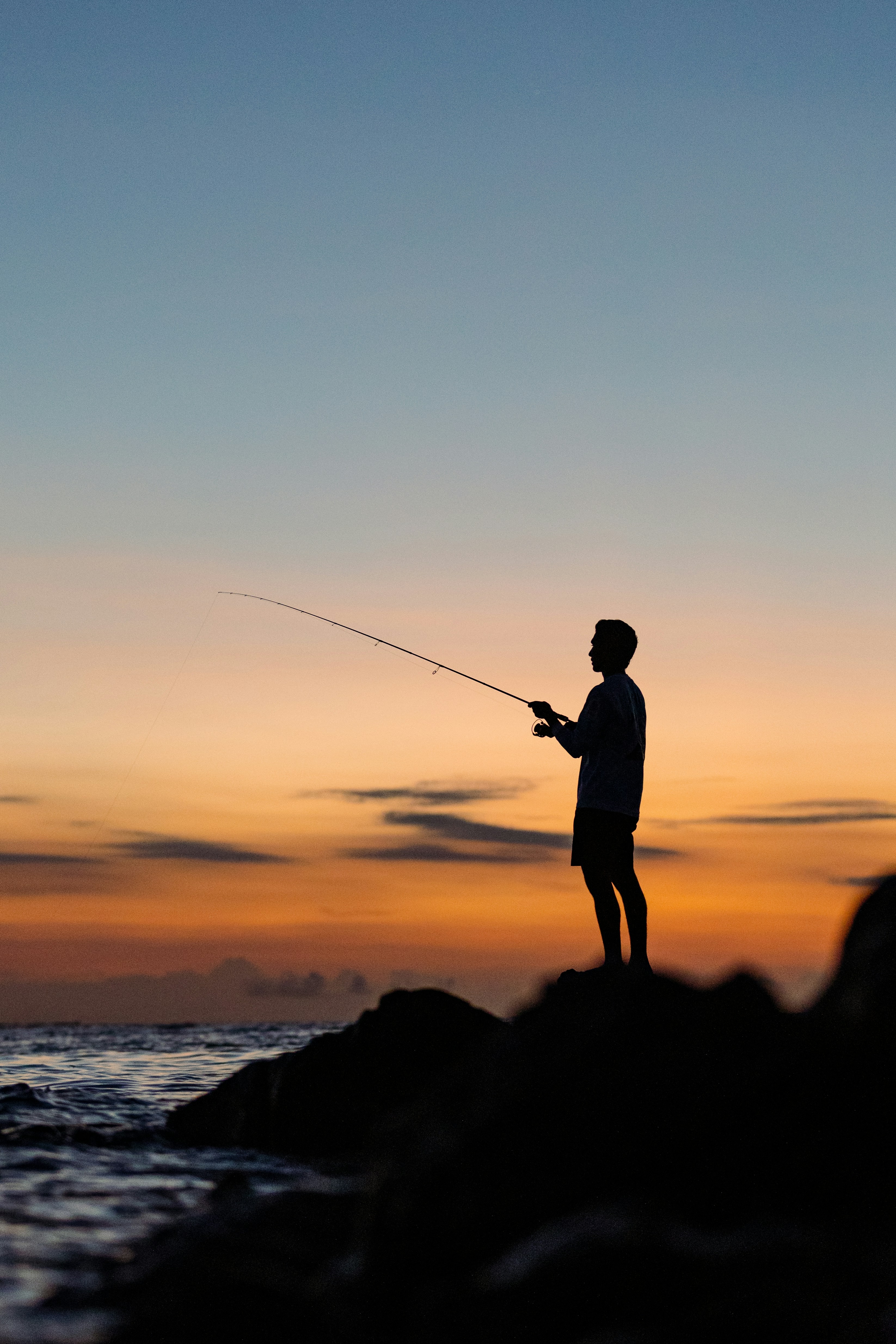 Photo of A person in silhouette fishing from a rocky shore against a warm orange sky by shaamil ali