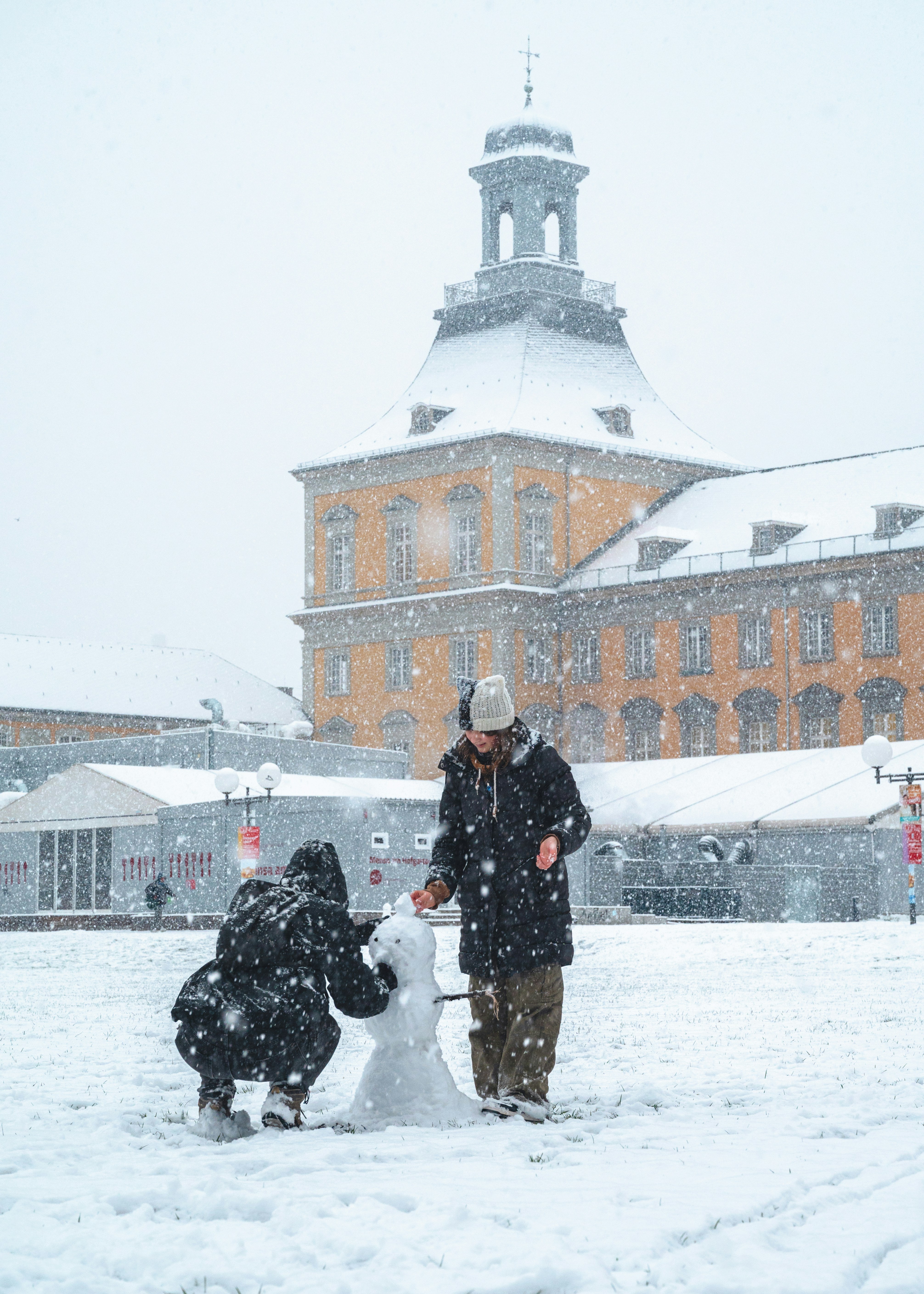 Dos personas construyendo un muñeco de nieve frente a un edificio