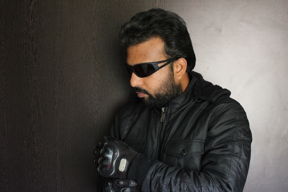 a man in a black jacket and sunglasses leaning against a wall