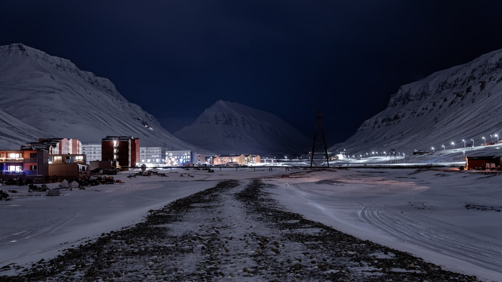 a snowy road at night with buildings and mountains in the background
