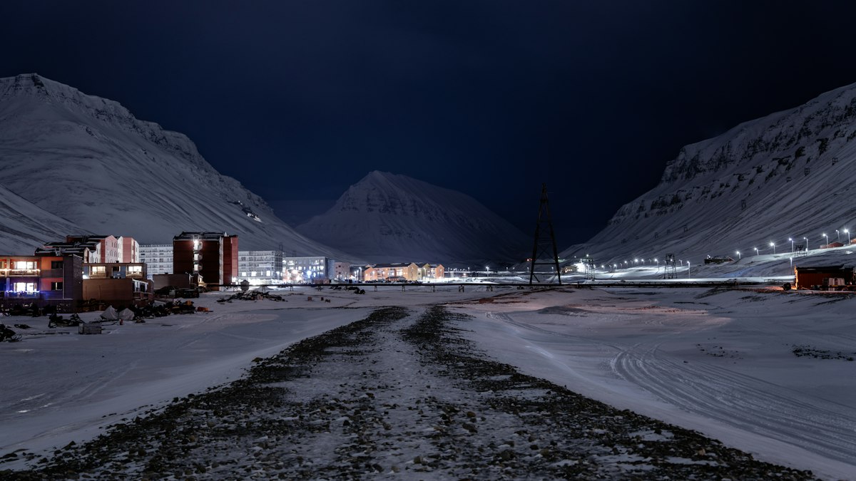 An Arctic town in darkness: a snowy gravel road leads to a row of lit, colourful apartment blocks, with snow-covered mountain slopes on both sides and a triangular snow-covered peak at the centre of the background under a blue-black sky. Street lamps burn along the road on the right