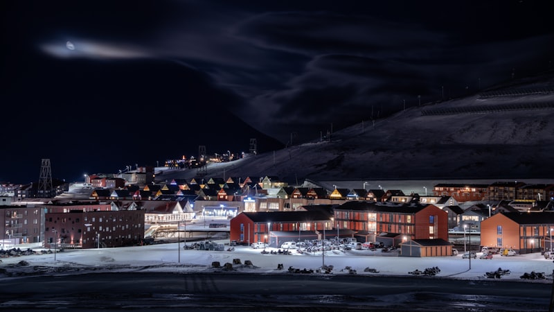 Snow-covered Arctic town at night with lit red and orange buildings, string lights on a hillside, and a dark mountain looming behind