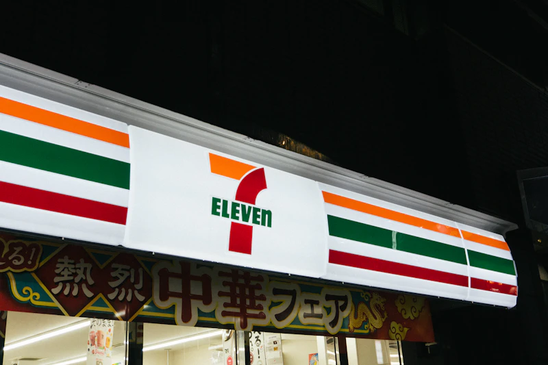 7-Eleven Japan store sign lit up at night with Japanese promotional banner visible below
