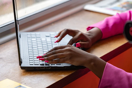 Close-up of hands typing on a laptop at a wooden desk by a window