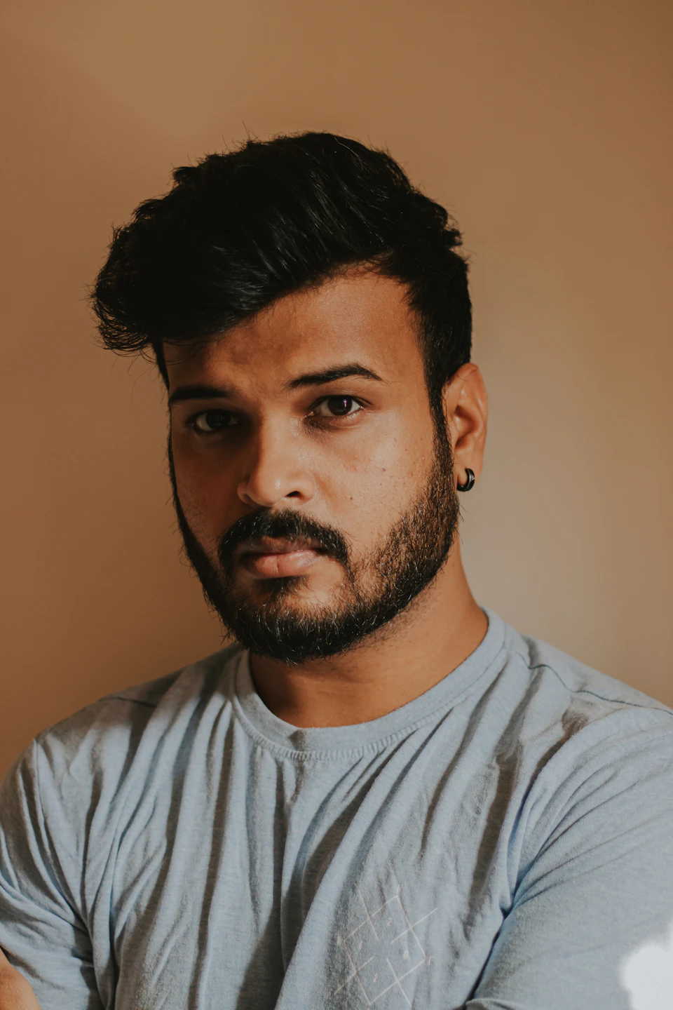 The image shows a man with dark hair and a beard wearing a light blue t-shirt against a beige background. The man's gaze is directed straight at the camera, creating an engaging portrait.