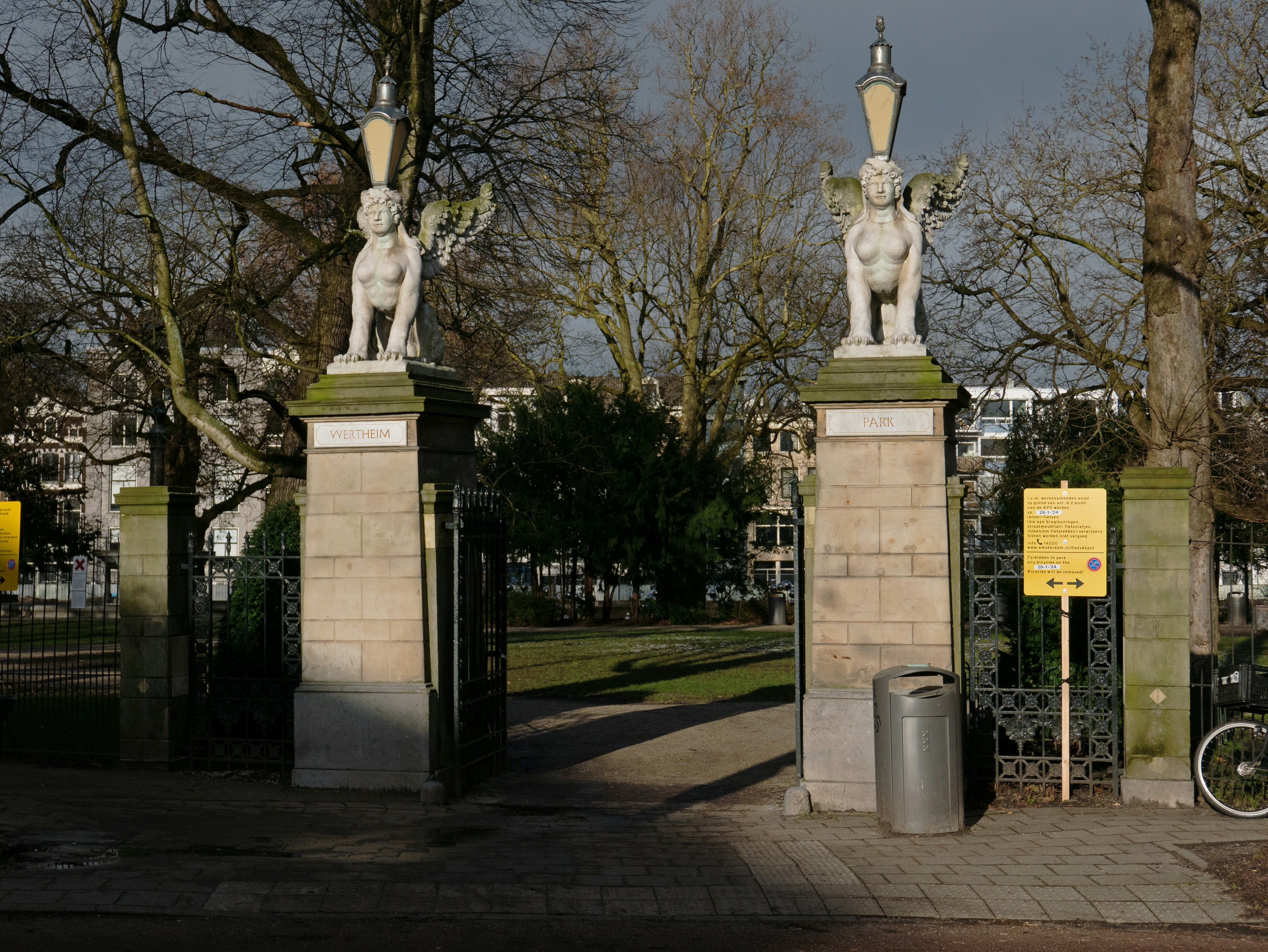 a couple of statues sitting on top of a sidewalk, Photo of two sfinxs sculptures on both sides of the entrance of the Wertheim park in Amsterdam city, Plantage district. On a sunny winterday with a lot of shades and cloudy sky. Nice combination! I like the tree branches with the grey background of the sky. Street photography of urban city parks in The Netherlands by Fons Heijnsbroek, 2024; Dutch photographer // Foto van Amsterdam Wertheimpark in de Plantage, gratis download, Nederland.