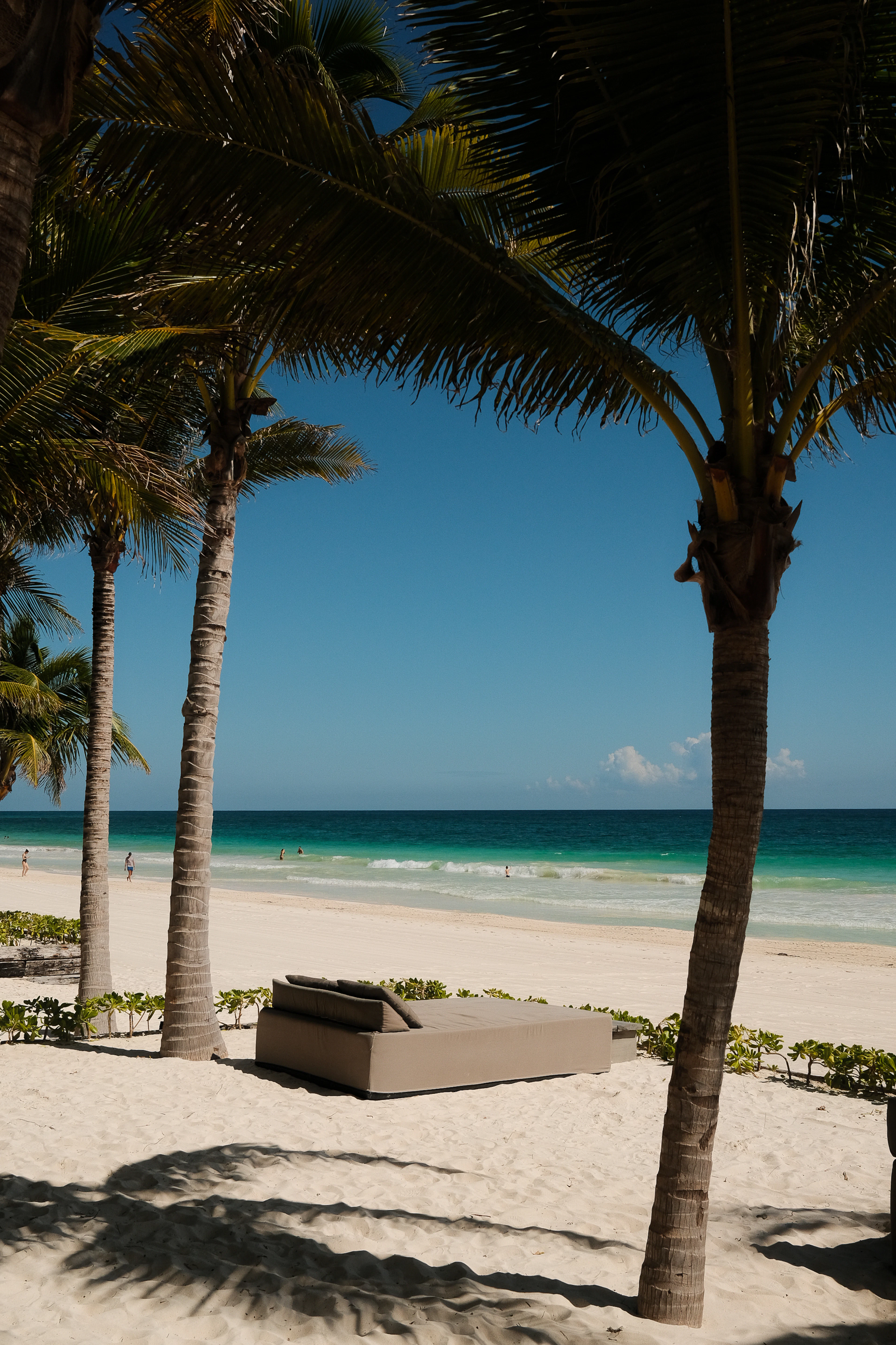 A cushioned daybed set out on the white sand between coconut palms on Tulum's hotel beach, the Caribbean shading from turquoise shallows to deep blue beyond the surf.